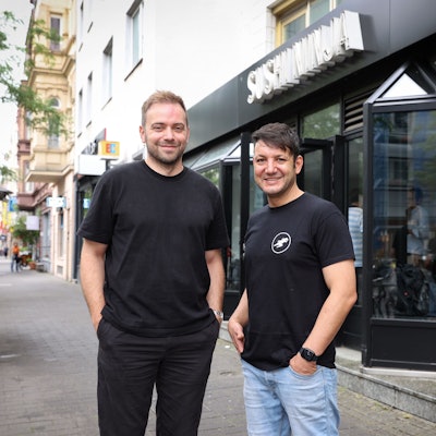 David Keuenhof und Sofien Kouraichi stehen vor einem von drei Kölner Sushi-Ninja-Restaurants. Sie tragen dunkle T-Shirts.