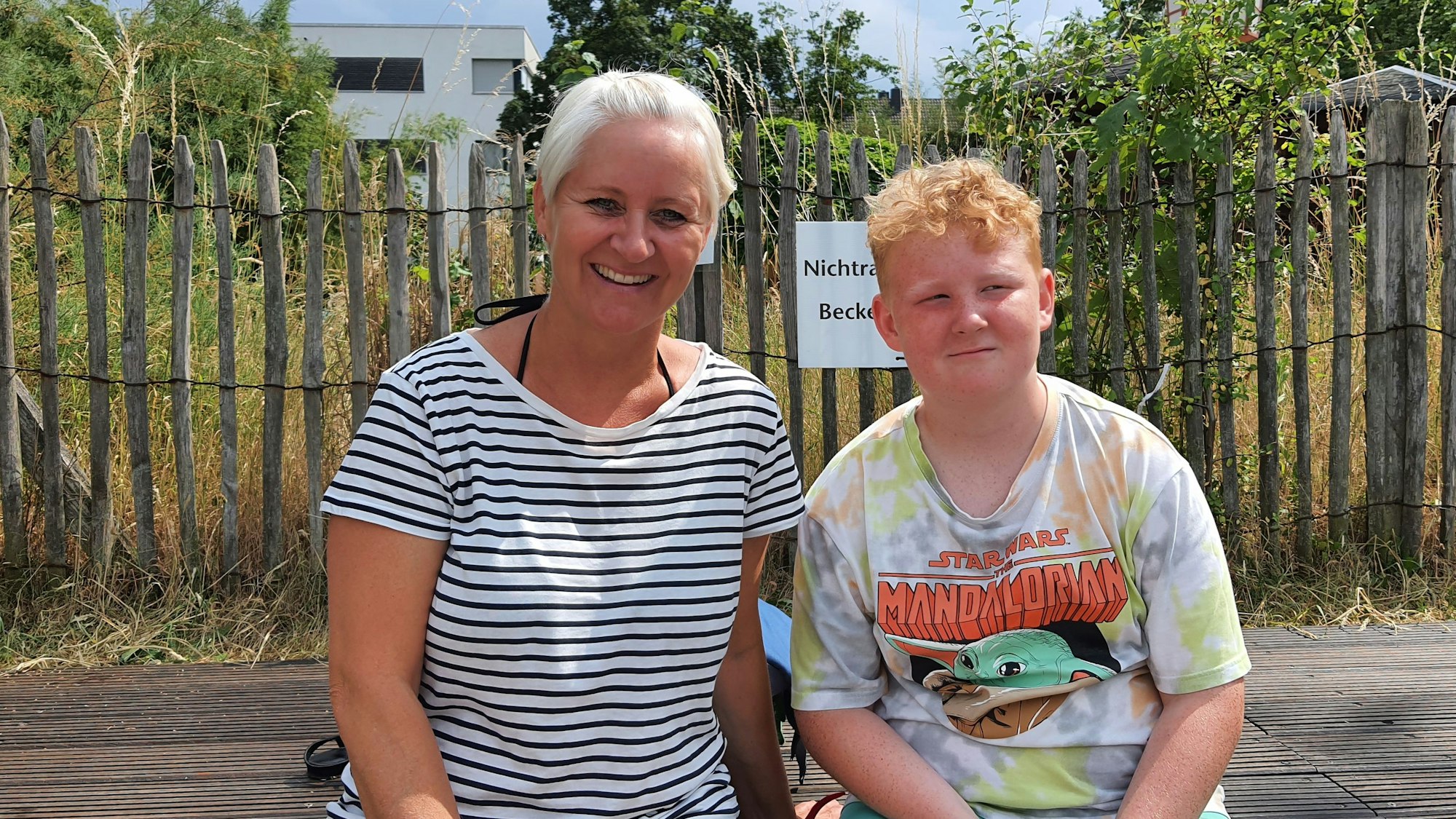 Eine Mutter und ihr Sohn sitzen vor einem Zaun und hohen Gräsern.