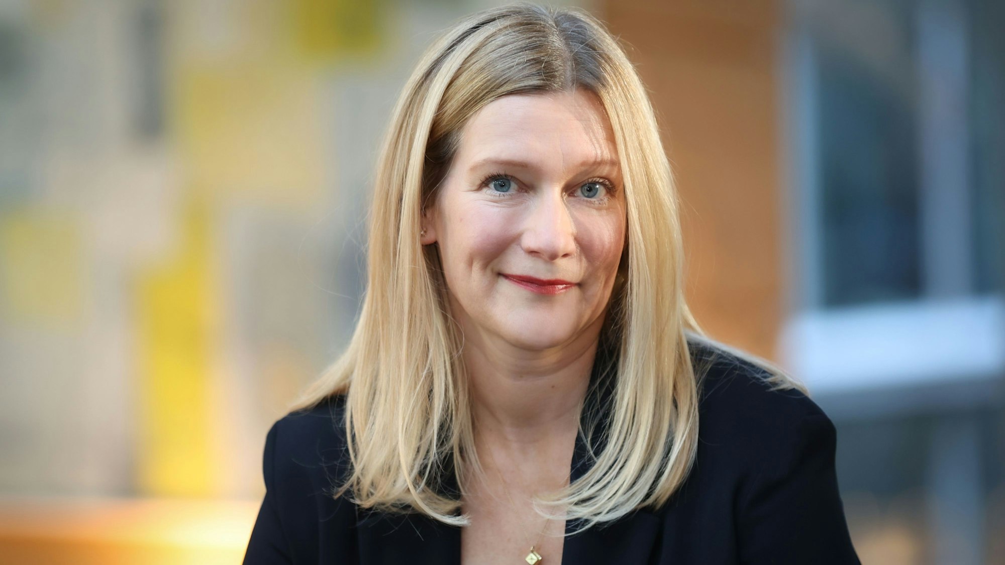 Portrait von Sabine Cramer, sie trägt ein schwarzes Sakko und eine goldene Kette. Sie hat lange, blonde Haare.