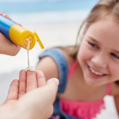 Eine Person macht Sonnencreme auf ihre Hand. Im Hintergrund ist ein lächelndes Mädchen zu sehen.