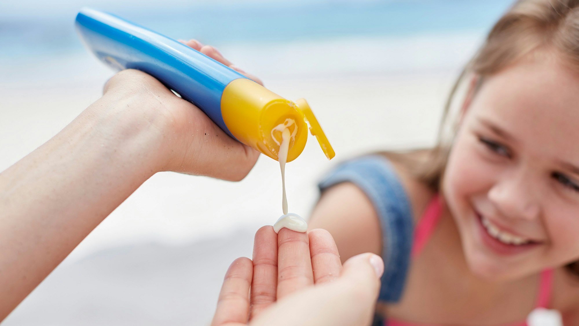 Eine Person macht Sonnencreme auf ihre Hand. Im Hintergrund ist ein lächelndes Mädchen zu sehen.