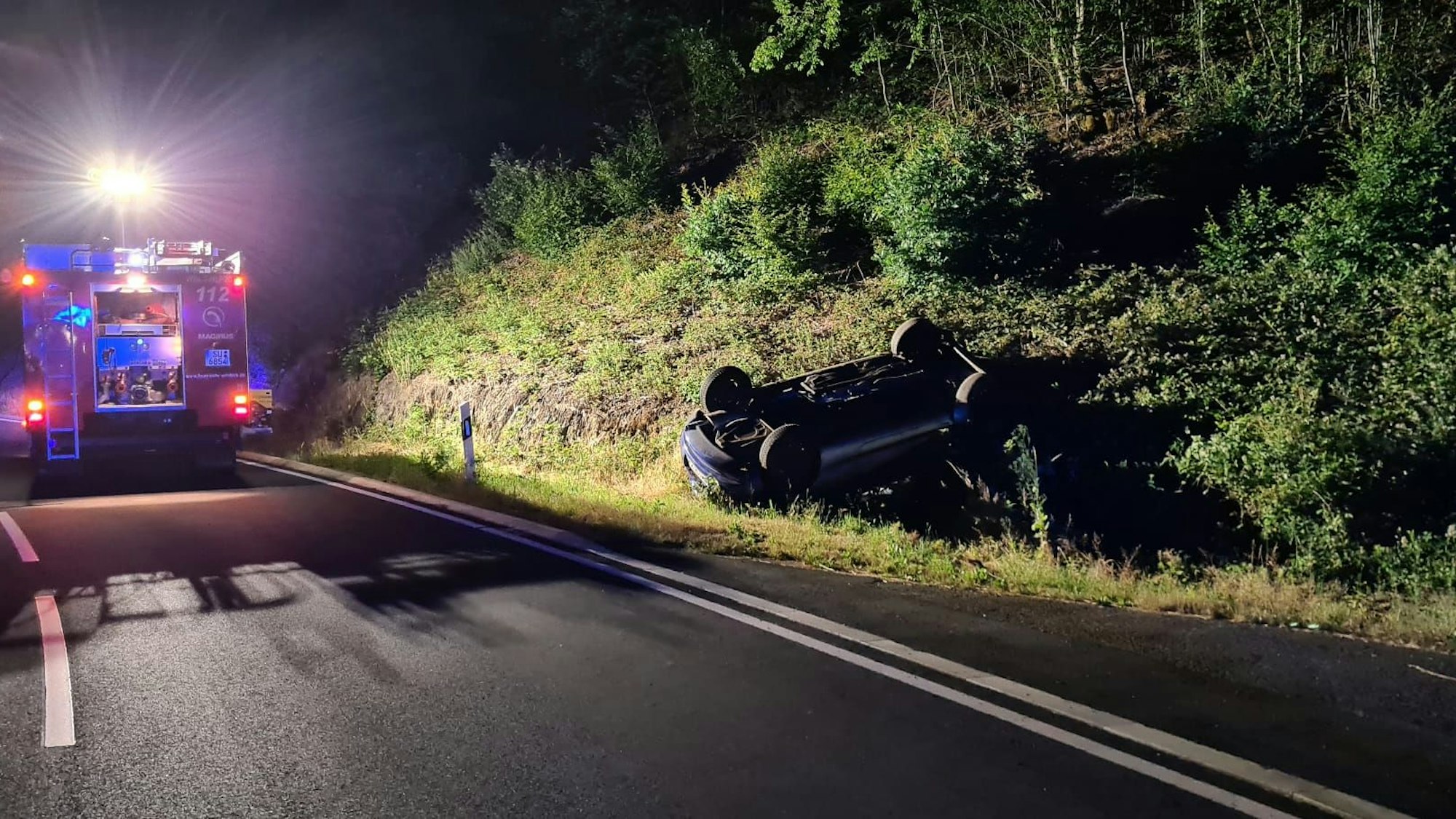 Im Graben neben einer Straße liegt ein Auto auf dem Kopf. Auf der Straße steht ein Feuerwehrauto.