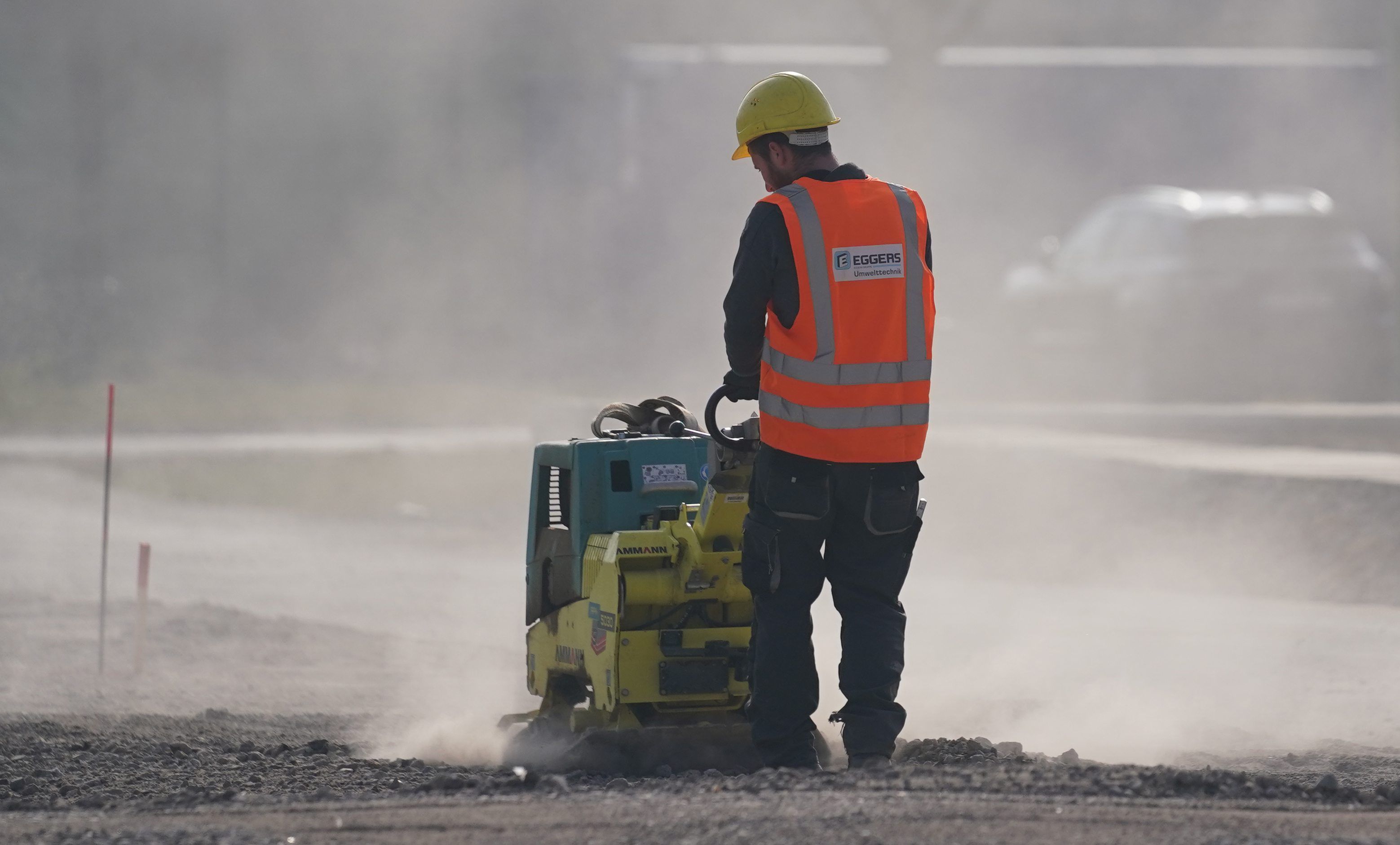 Ein Arbeiter ist auf einer Baustelle zu sehen. Er bedient ein Gerät, es staubt, er trägt einen gelben Helm und eine orange Warnweste.