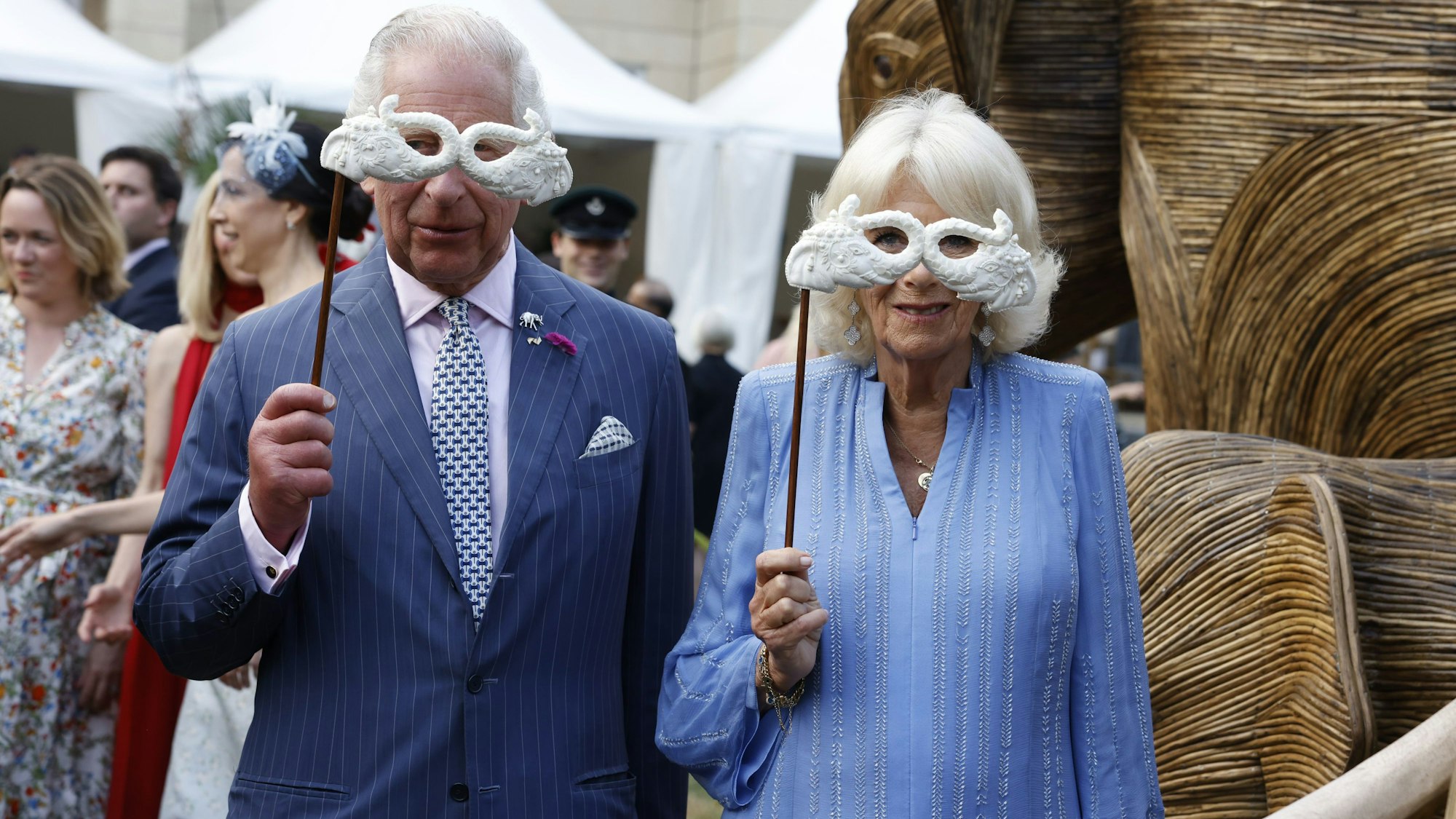 König Charles III. und Königin Camilla halten sich lustige Masken an Holzstäben vor das Gesicht.