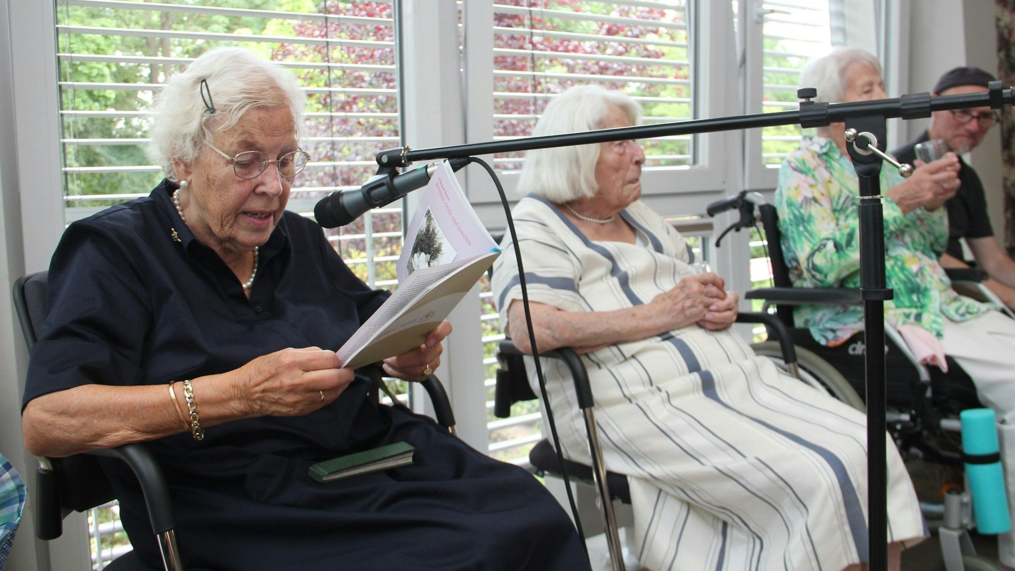 Drei ältere Damen sitzen nebeneinander, eine liest aus einem Buch, sie spricht in ein Mikrofon.