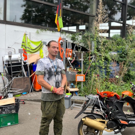 Der Obdachlose Marcel K. steht vor seiner Hütte am Höhenbergbad. Neben ihm ein Motorroller.