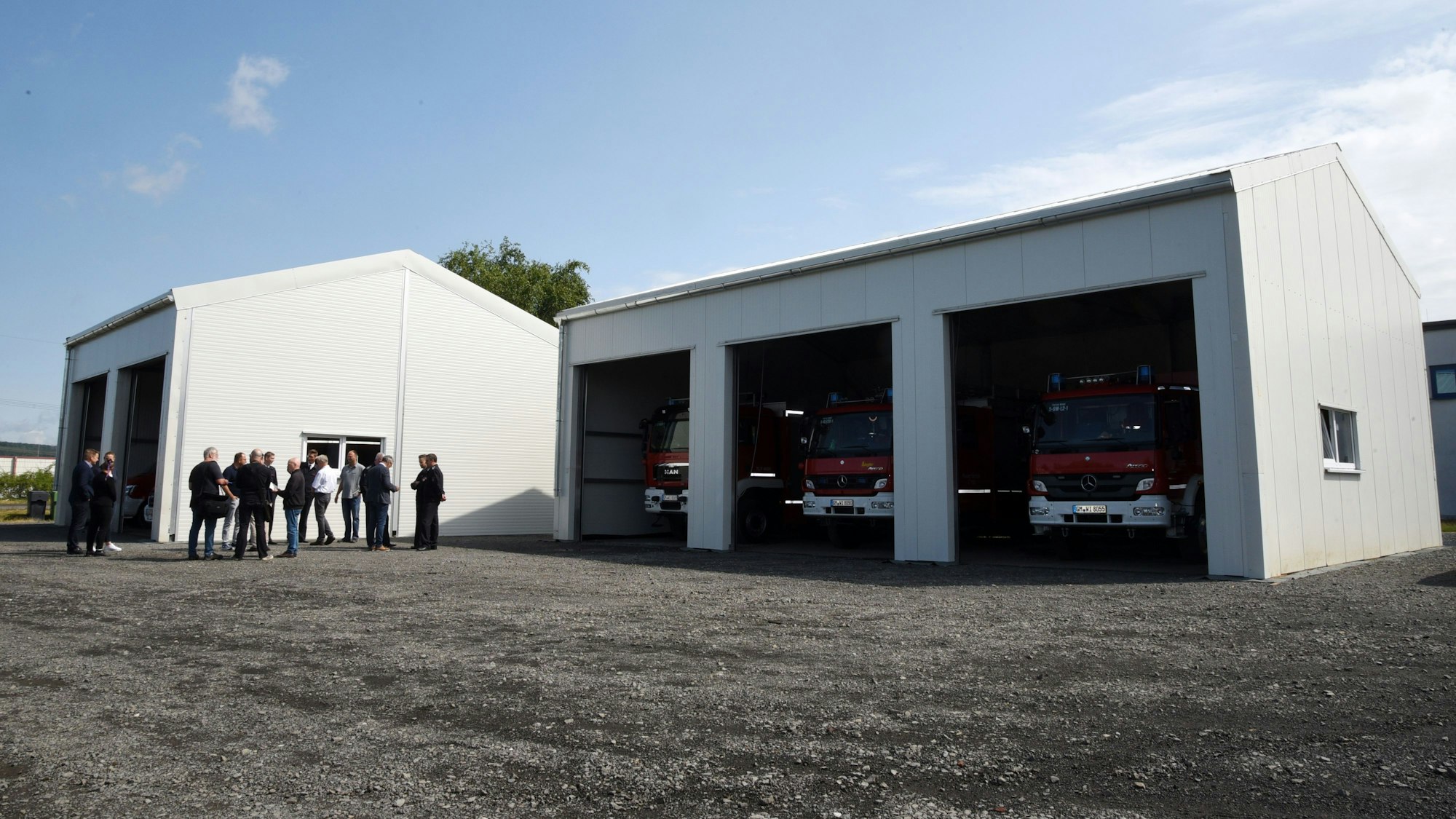 Eine Gruppe Menschen steht vor zwei großen weißen Hallen, in der rechten sind drei Feuerwehrautos geparkt.