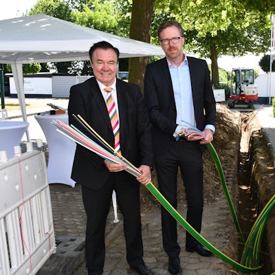 Zwei Männer mit Kabelenden in den Händen vor einer Baugrube.
