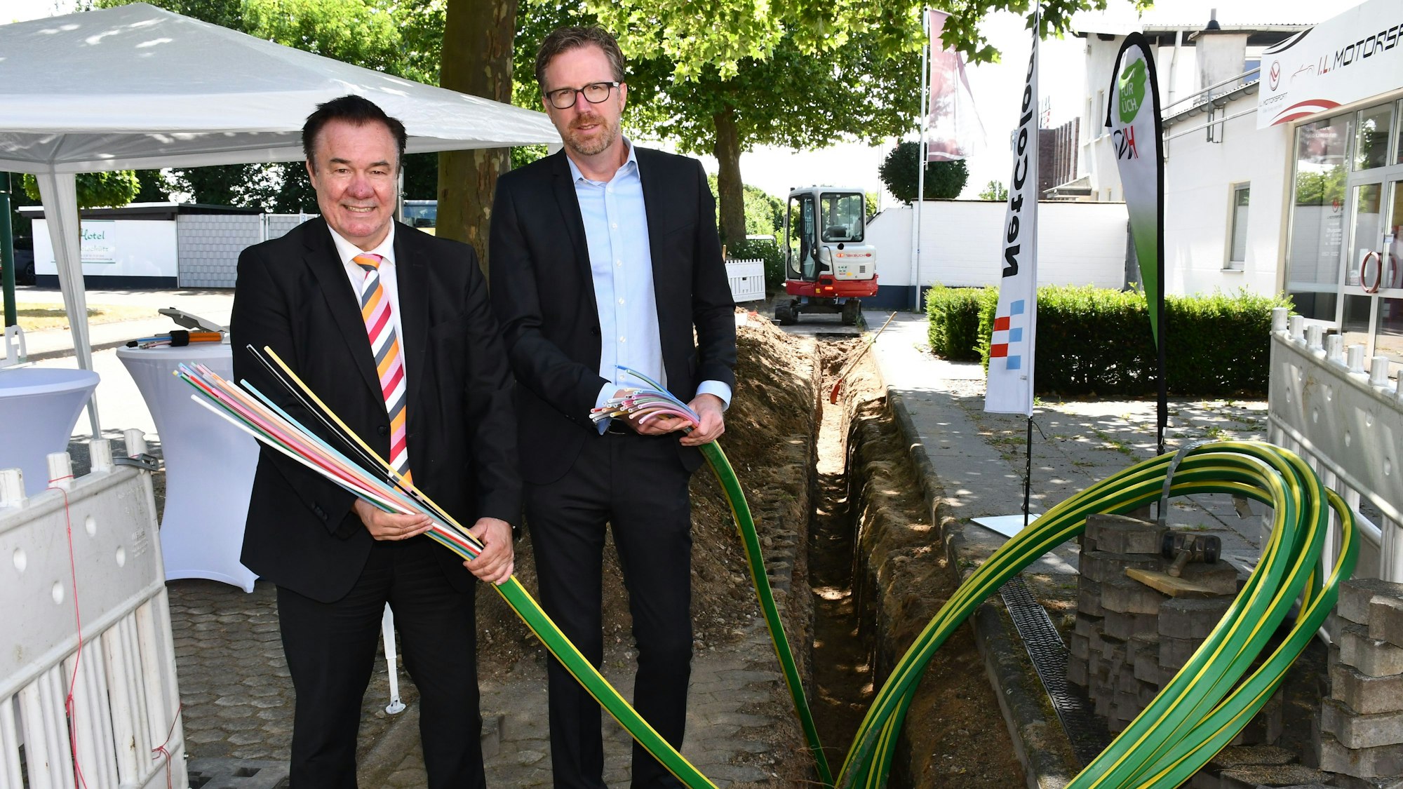 Zwei Männer mit Kabelenden in den Händen vor einer Baugrube.