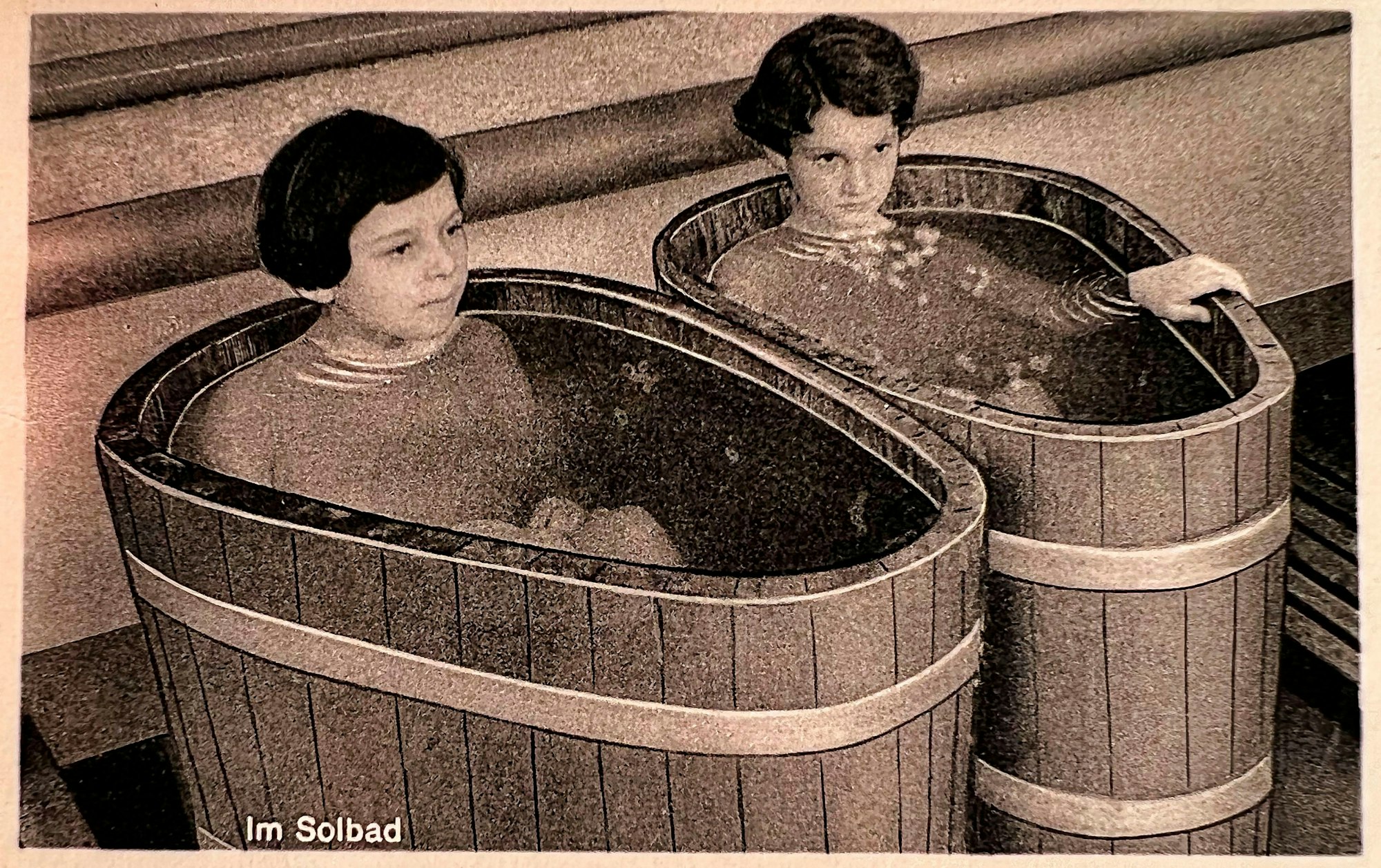 Zwei kleine Holzbadewannen stehen nebeneinander; in ihnen sitzt jeweils ein Kind. Ihre Körper sind bis zum Hals von Solewasser bedeckt. Das Foto ist alt und vergilbt und trägt die Aufschrift „Im Solebad“.