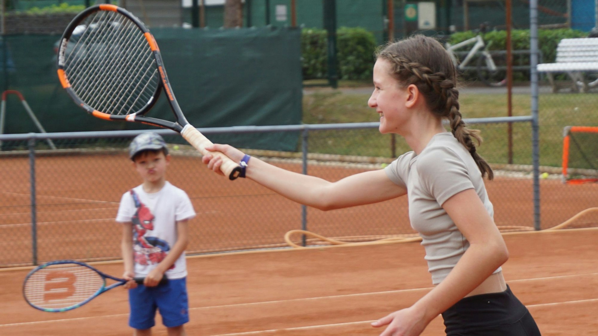 Ein junges Mädchen steht auf dem Tennisplatz und hält einen Tennisschläger in Erwartung eines Balles in die Luft. Ein Junge mit einem Schläger in der Hand sieht ihr zu.