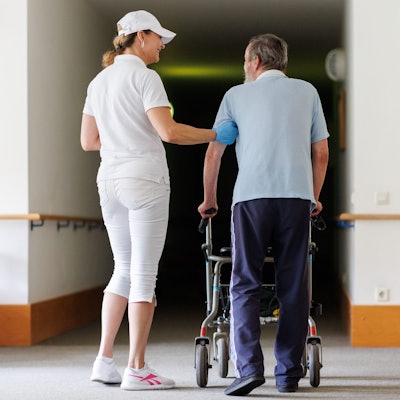 Svetlana (l.), ambulante Pflegefachkraft, begleitet einen Bewohner des Betreuten Wohnens mit seiner Gehhilfe durch das Gebäude. (Symbolbild)