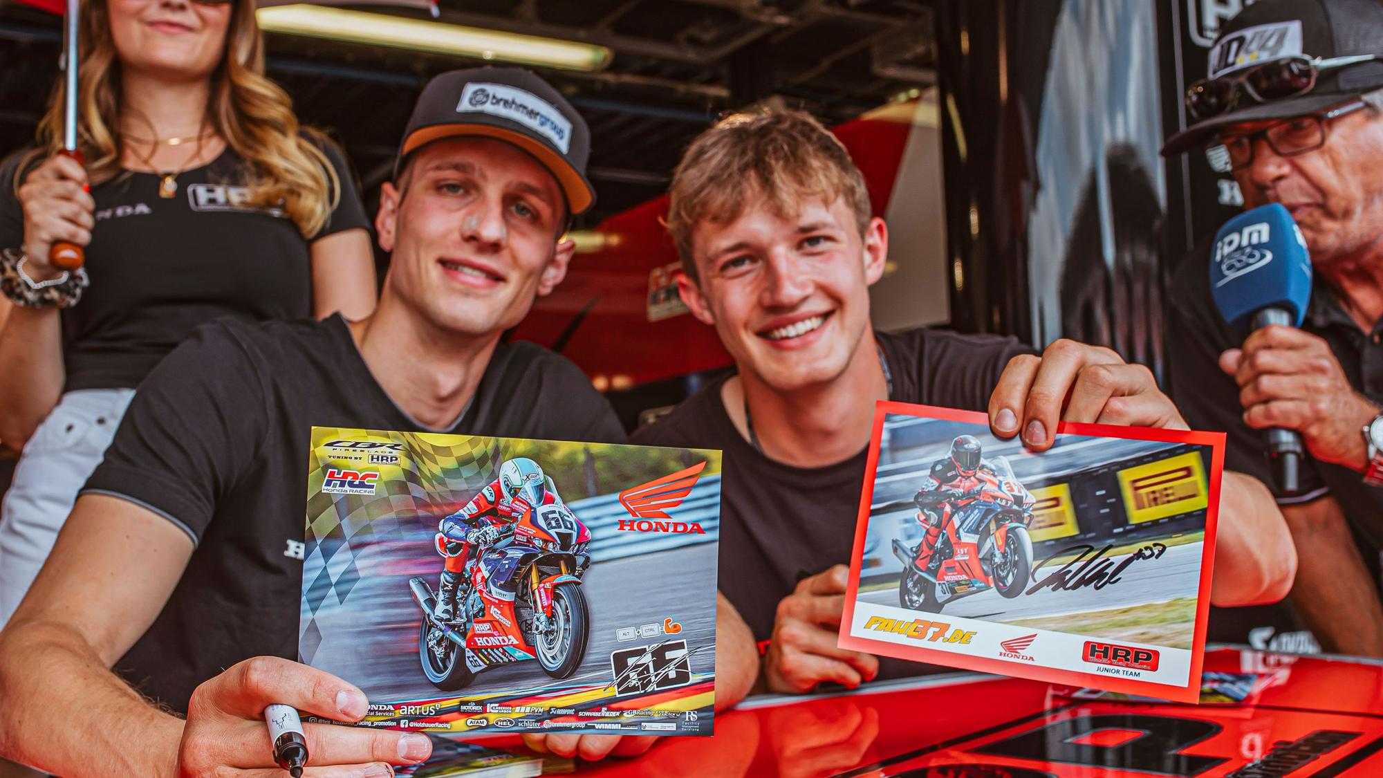 Zwei Motorradrennfahrer signieren Autogrammkarten.