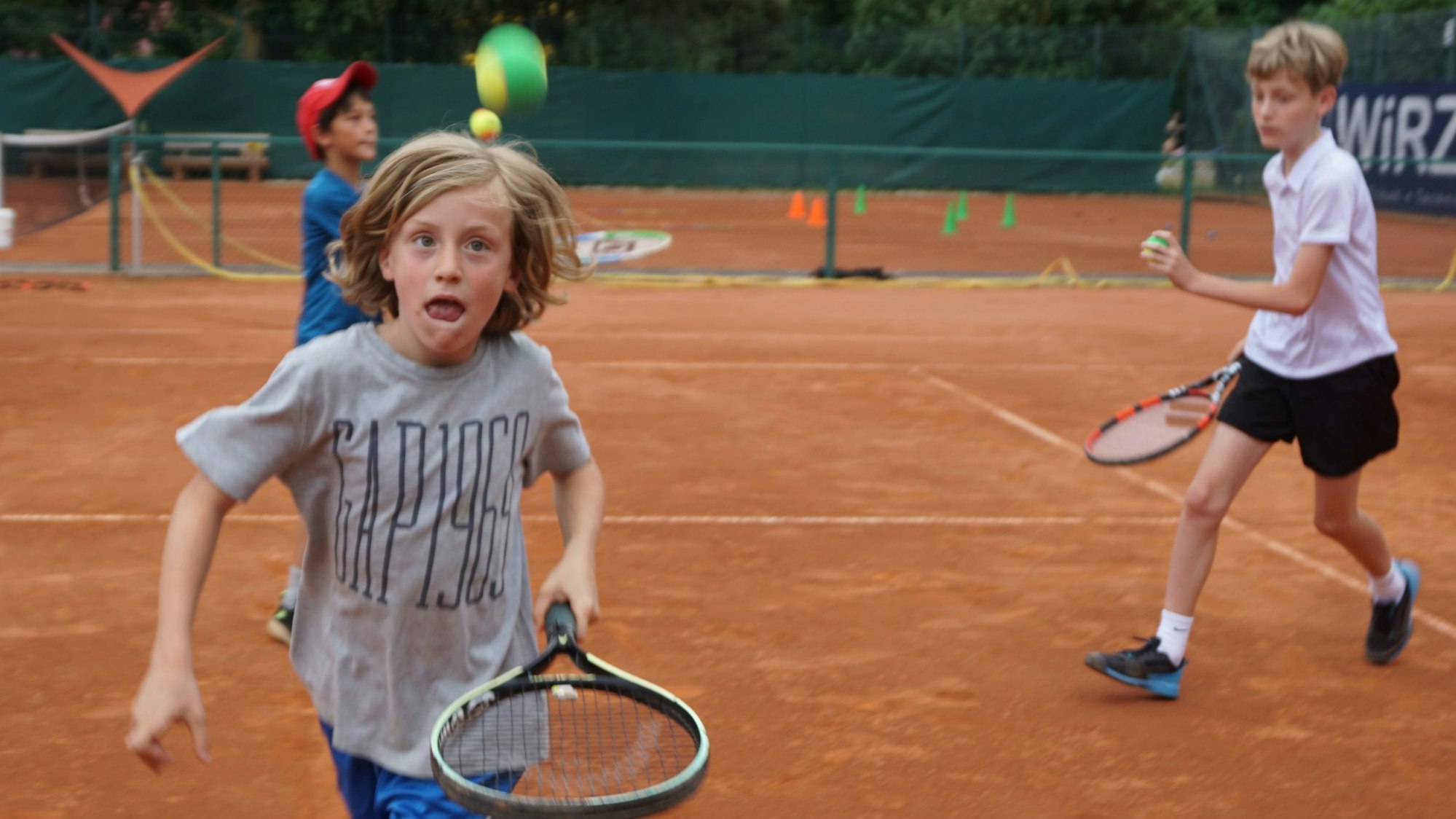 Ein Junge läuft mit einem Schläger über den Tennisplatz. Ein Tennisball fliegt durch die Luft. Der Junge hat einen konzentrierten Gesichtsausdruck.