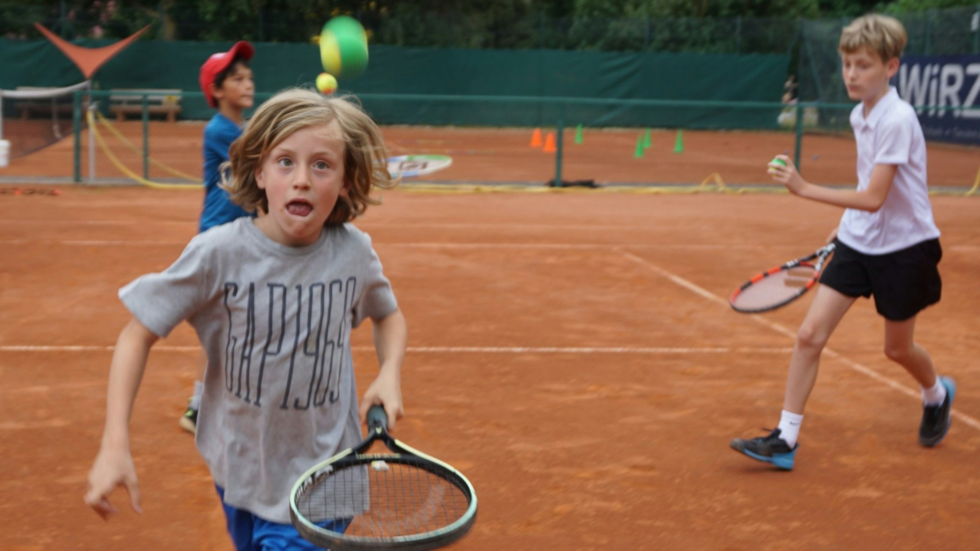 Ein Junge läuft mit einem Schläger über den Tennisplatz. Ein Tennisball fliegt durch die Luft. Der Junge hat einen konzentrierten Gesichtsausdruck.