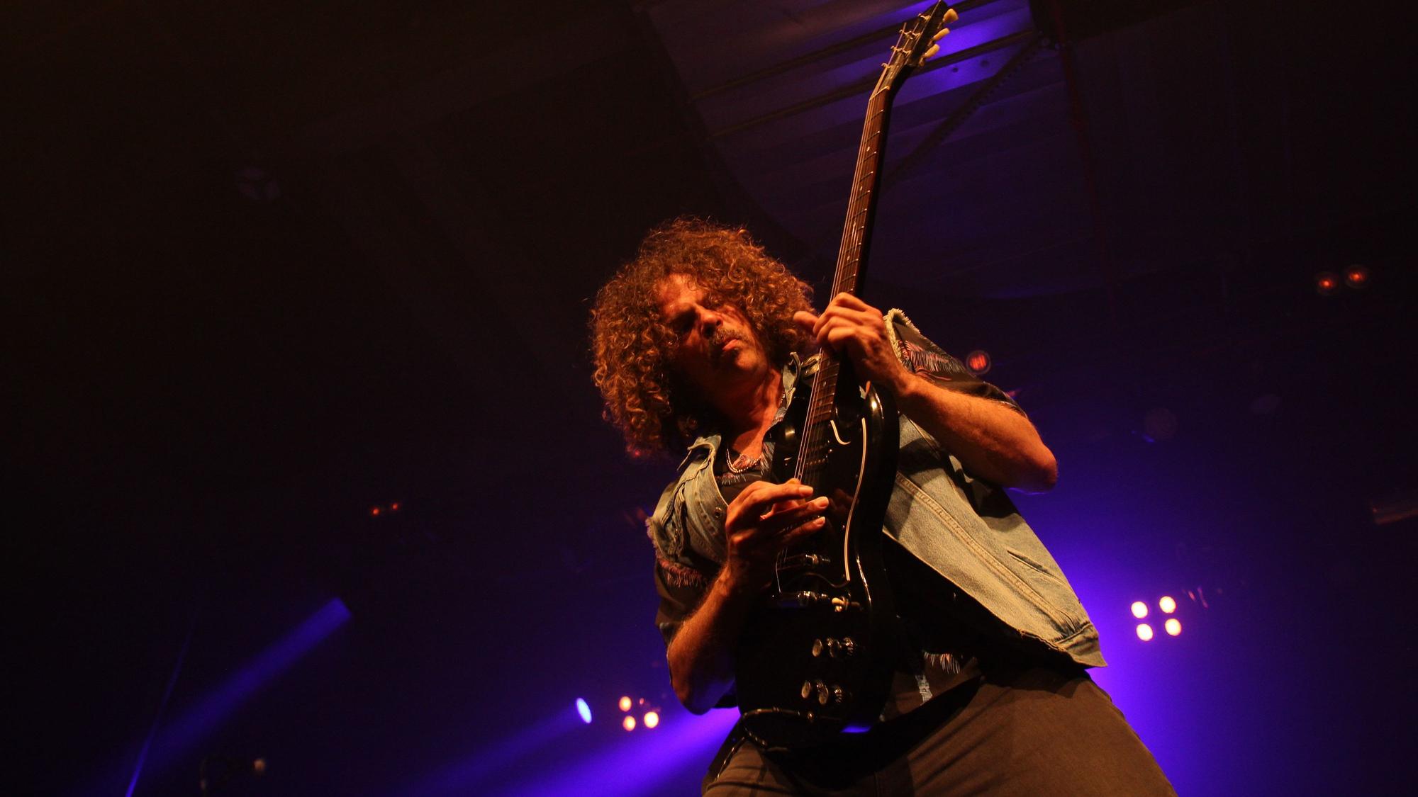 Andrew Stockdale von Wolfmother ist in sein Gitarrenspiel vertieft. Im Hintergrund glimmen die Bühnenscheinwerfer.