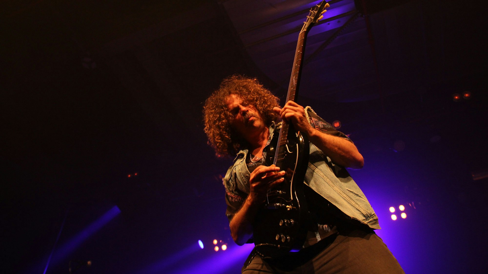 Andrew Stockdale von Wolfmother ist in sein Gitarrenspiel vertieft. Im Hintergrund glimmen die Bühnenscheinwerfer.