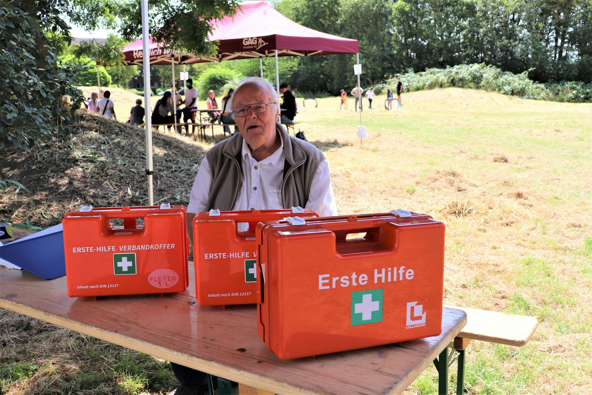 Ein älterer Herr sitzt an einem Biertisch, auf dem drei Erste-Hilfe-Koffer stehen.