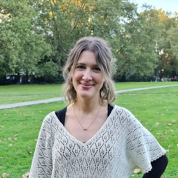 Eine junge Frau mit blondem kinnlangem Haar steht in einem Park.