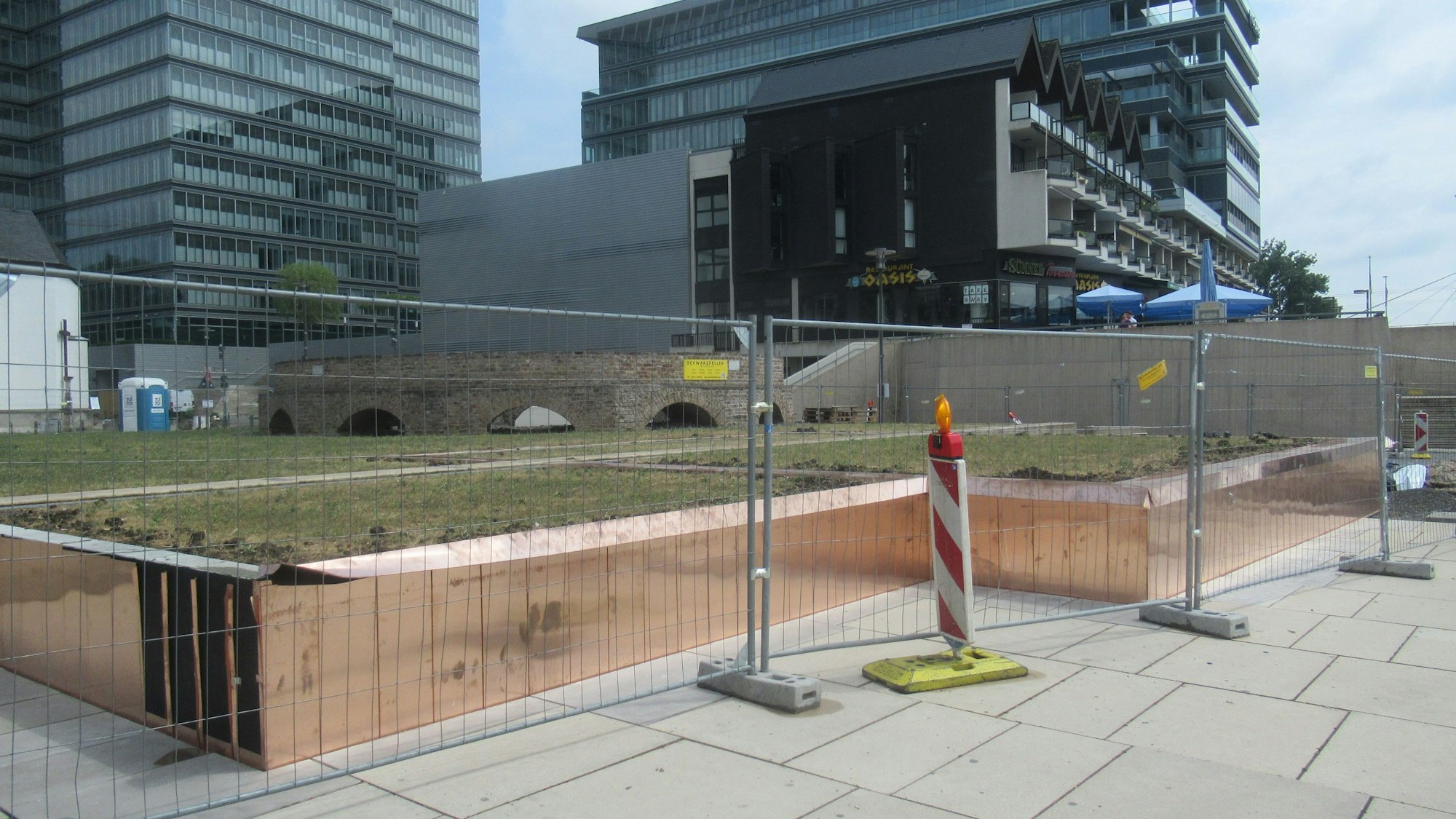 Hinter einem Bauzaun ist eine mit kupferfarbenem Metall eingefasste Rasenfläche zu sehen. Sie entspricht dem Grundriss der Pfarrkirche St. Heribert aus dem 11. Jahrhundert.