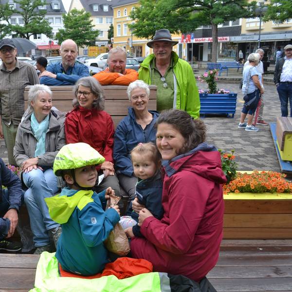 Erwachsene und Kinder in bunten Regenjacken sitzen auf Bänken und Blumenkästen aus Holz. Alle lachen.