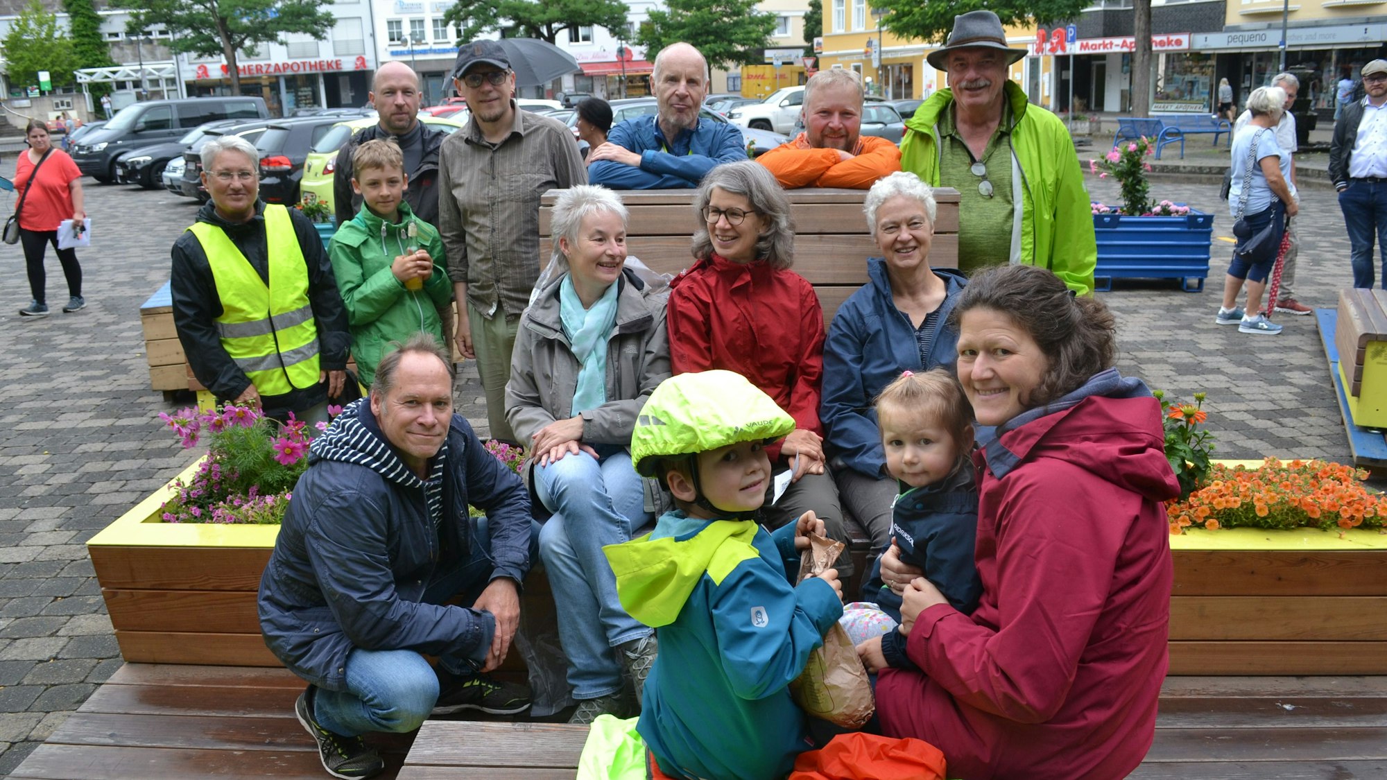 Erwachsene und Kinder in bunten Regenjacken sitzen auf Bänken und Blumenkästen aus Holz. Alle lachen.