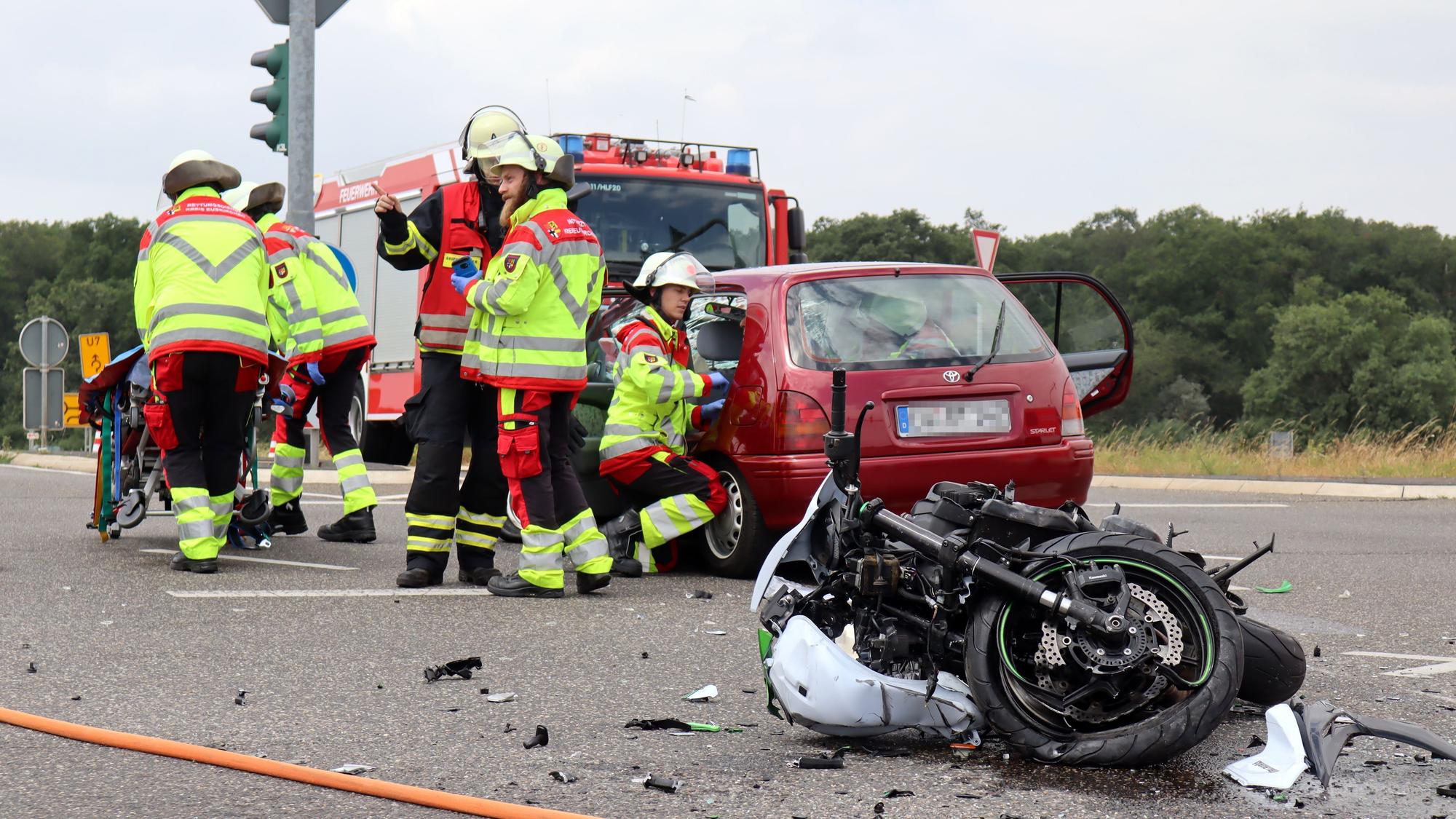 Ein schwer beschädigtes Motorrad liegt auf der Fahrbahn, dahinter stehen das an dem Unfall beteiligte Auto und ein Feuerwehrfahrzeug. Der Rettungsdienst kümmert sich um Verletzte.