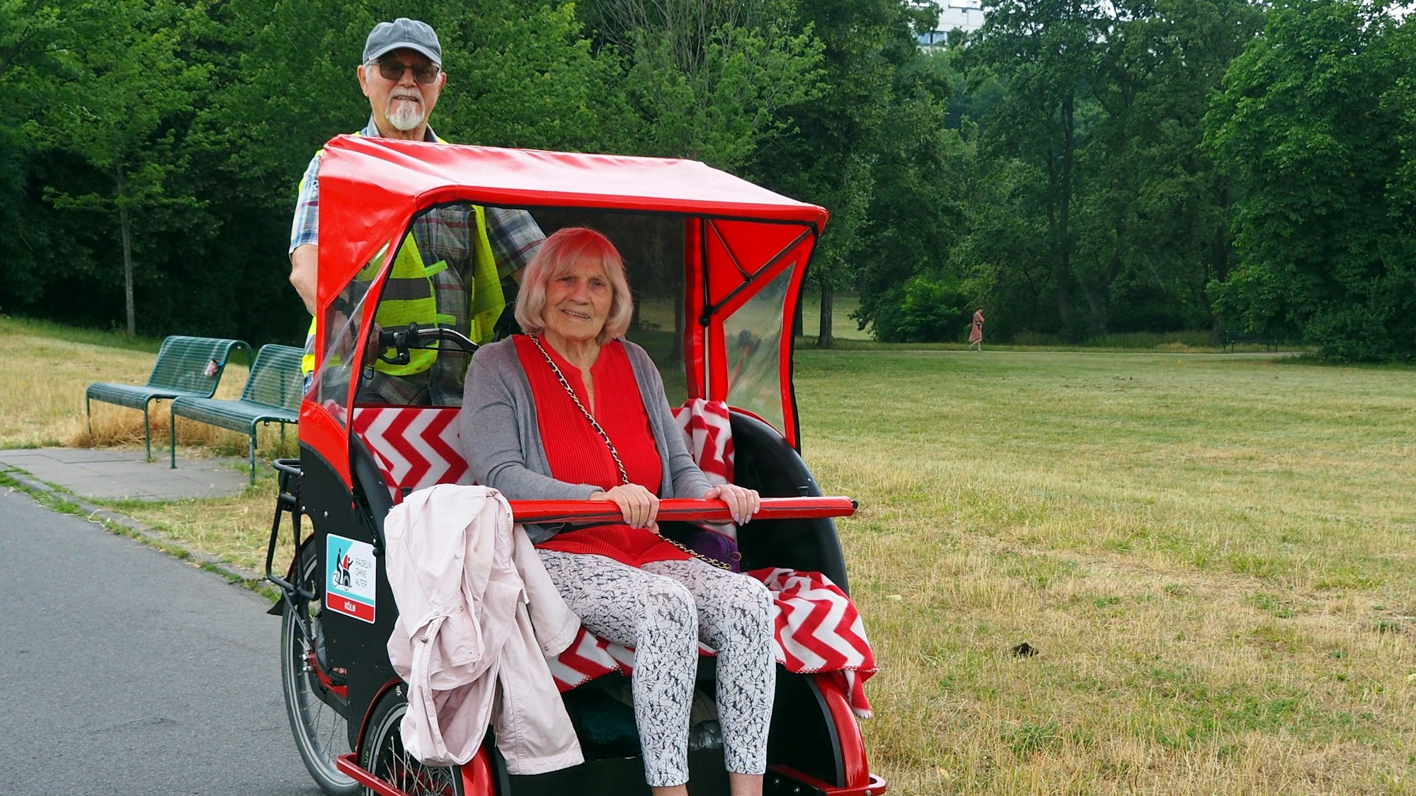 Eine ältere Dame sitzt in einer Rikscha, die ein Herr mit Bart und Schirmkappe fährt.