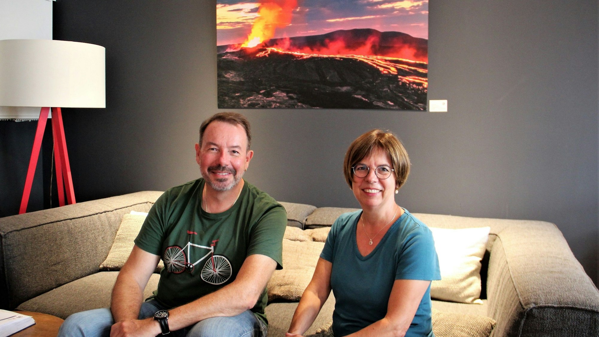 Ein Mann mit Bart und eine Frau mit Pagenkopf und Brille sitzen auf einem Sofa, hinter sich ist ein Bild von einem Vulkan-Ausbruch zu sehen.
