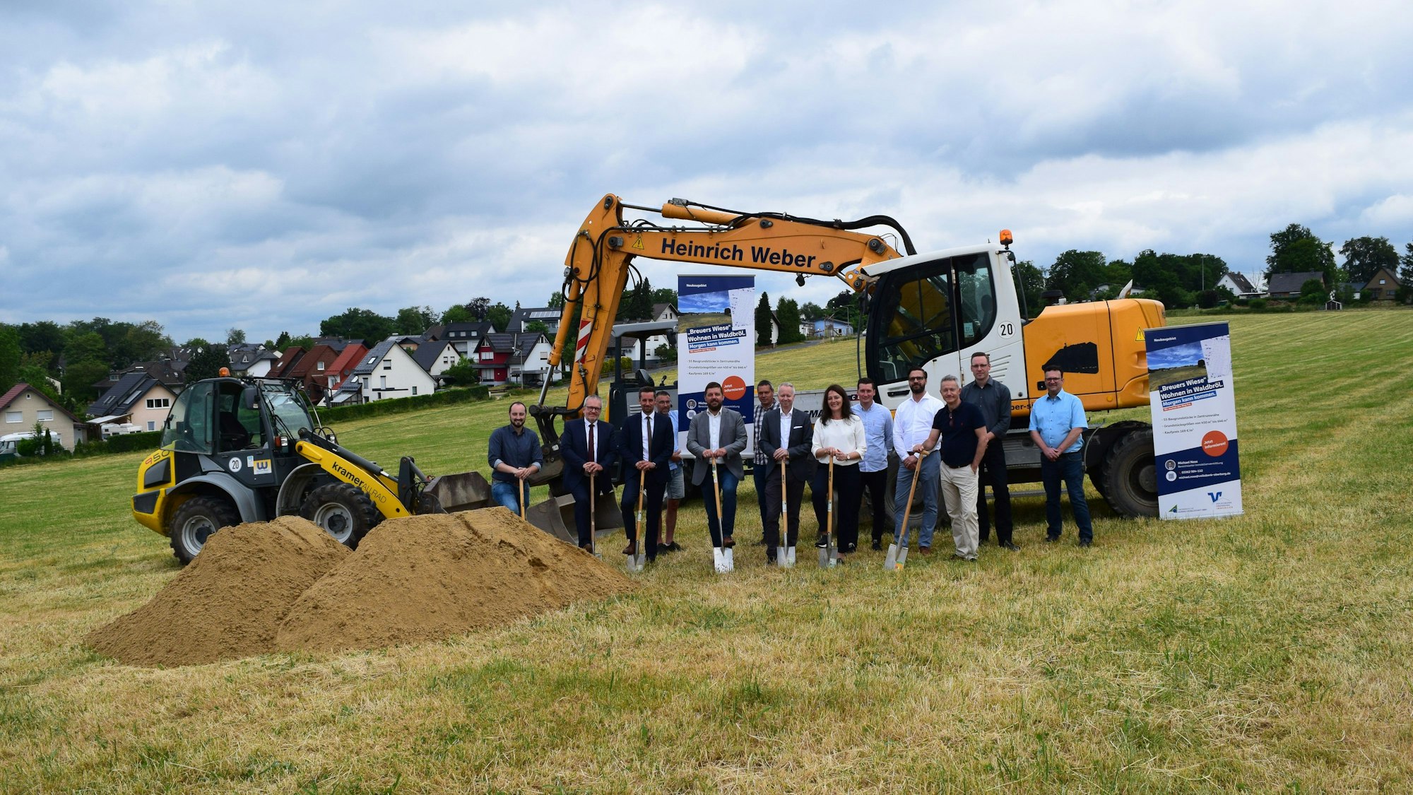 Ein Feld, auf dem mehrere Personen mit Spaten stehen. Im Vordergrund Sandhügel, hinter den Personen sind zwei Bagger zu sehen. Im Hintergrund sind Hausdächer zu erkennen.