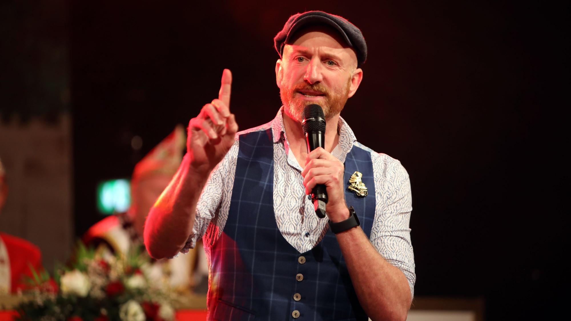 Martin Schops hält ein Mikrofon an seinen Mund und hat mit der anderen Hand einen Finger gehoben.