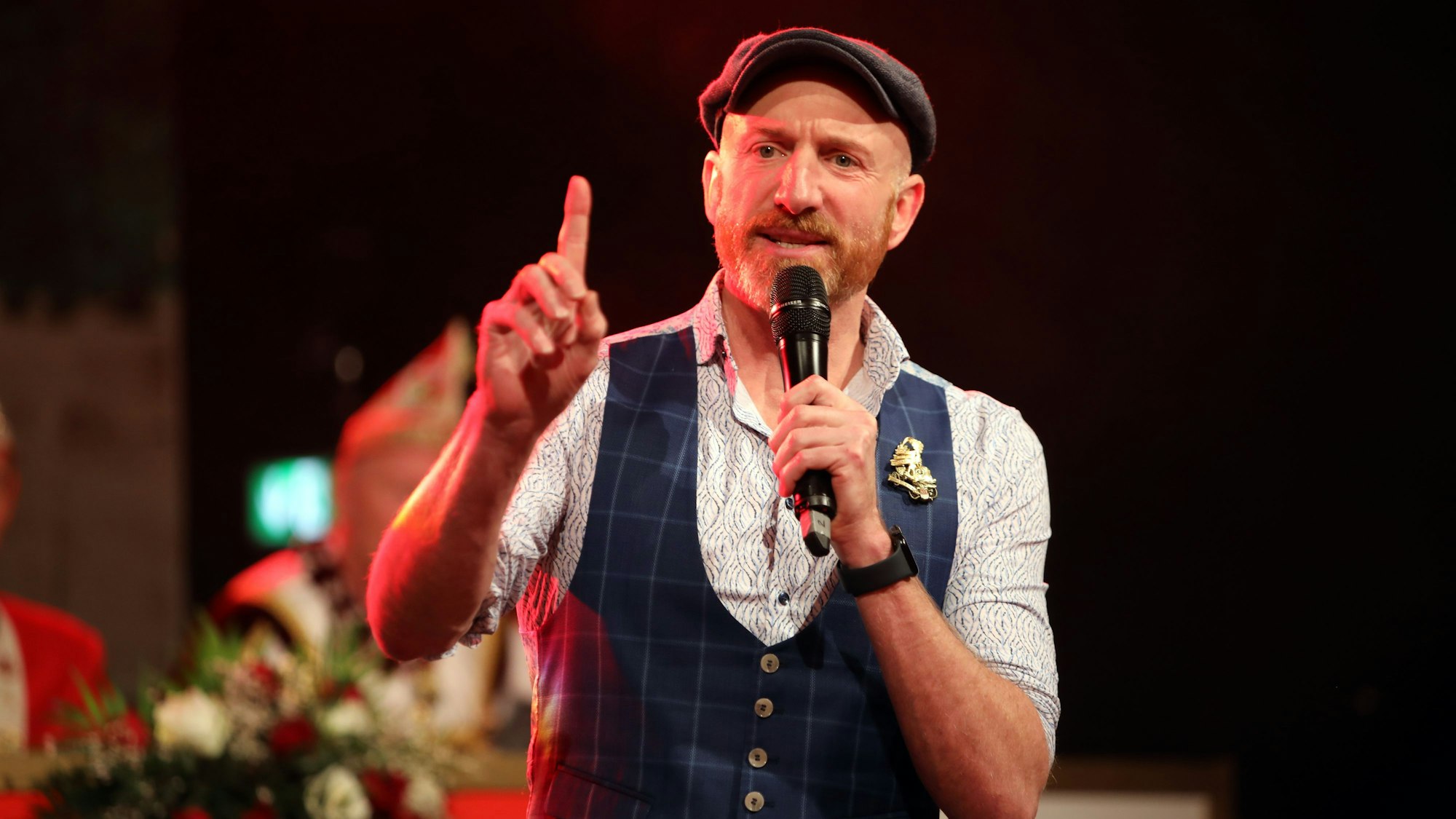 Martin Schops hält ein Mikrofon an seinen Mund und hat mit der anderen Hand einen Finger gehoben.