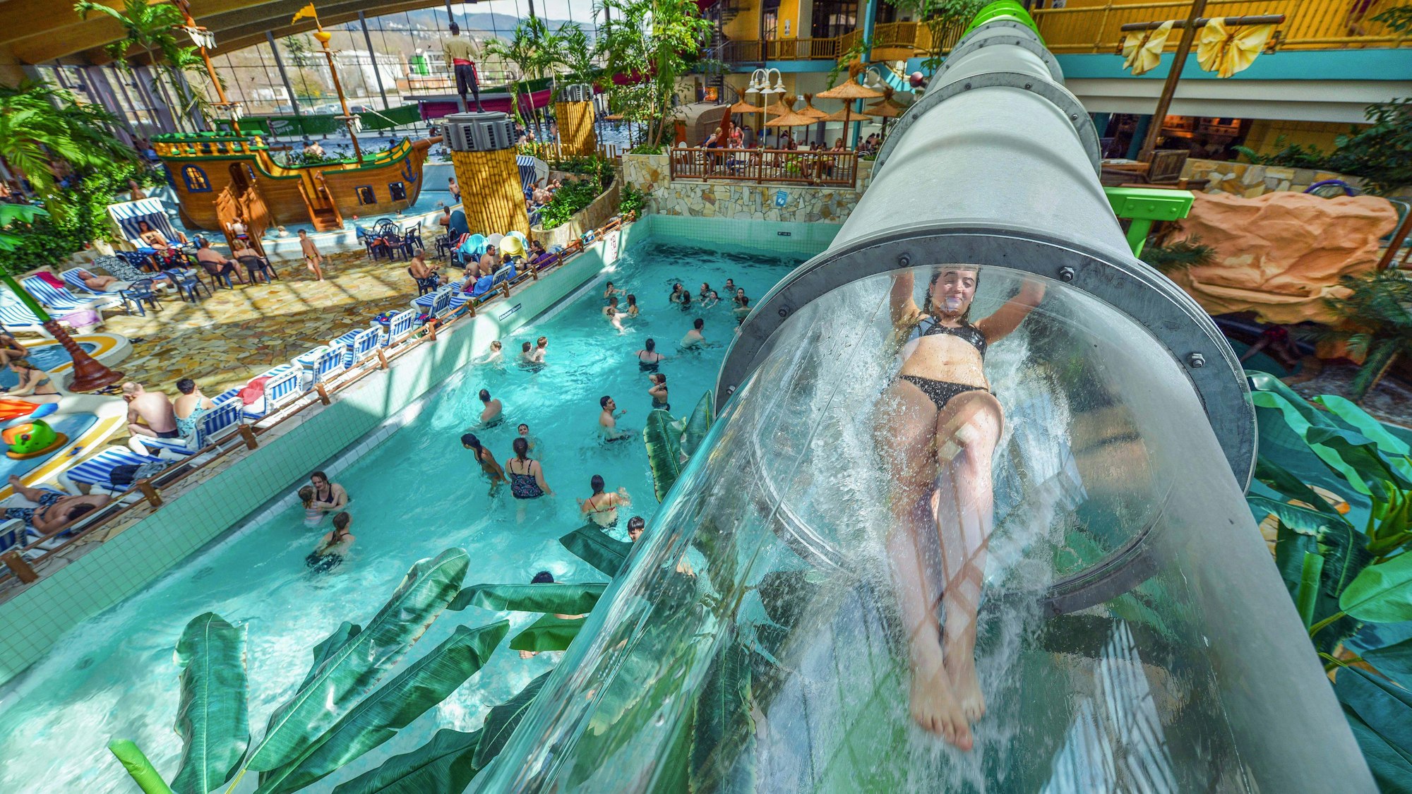 Frau rutscht durch eine durchsichtige Wasserrutsche, unter ihr Menschen in einem Schwimmbecken.