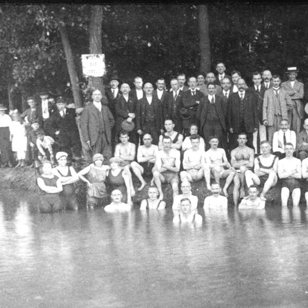 Auf einer Schwarz-Weiß-Fotografie sind viele Männer im Anzu zu sehen. Vor ihnen sitzen junge Leute am Ufer eines Sees, zum Teil im Wasser.