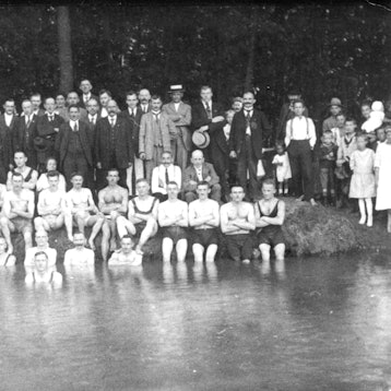 Auf einer Schwarz-Weiß-Fotografie sind viele Männer im Anzu zu sehen. Vor ihnen sitzen junge Leute am Ufer eines Sees, zum Teil im Wasser.
