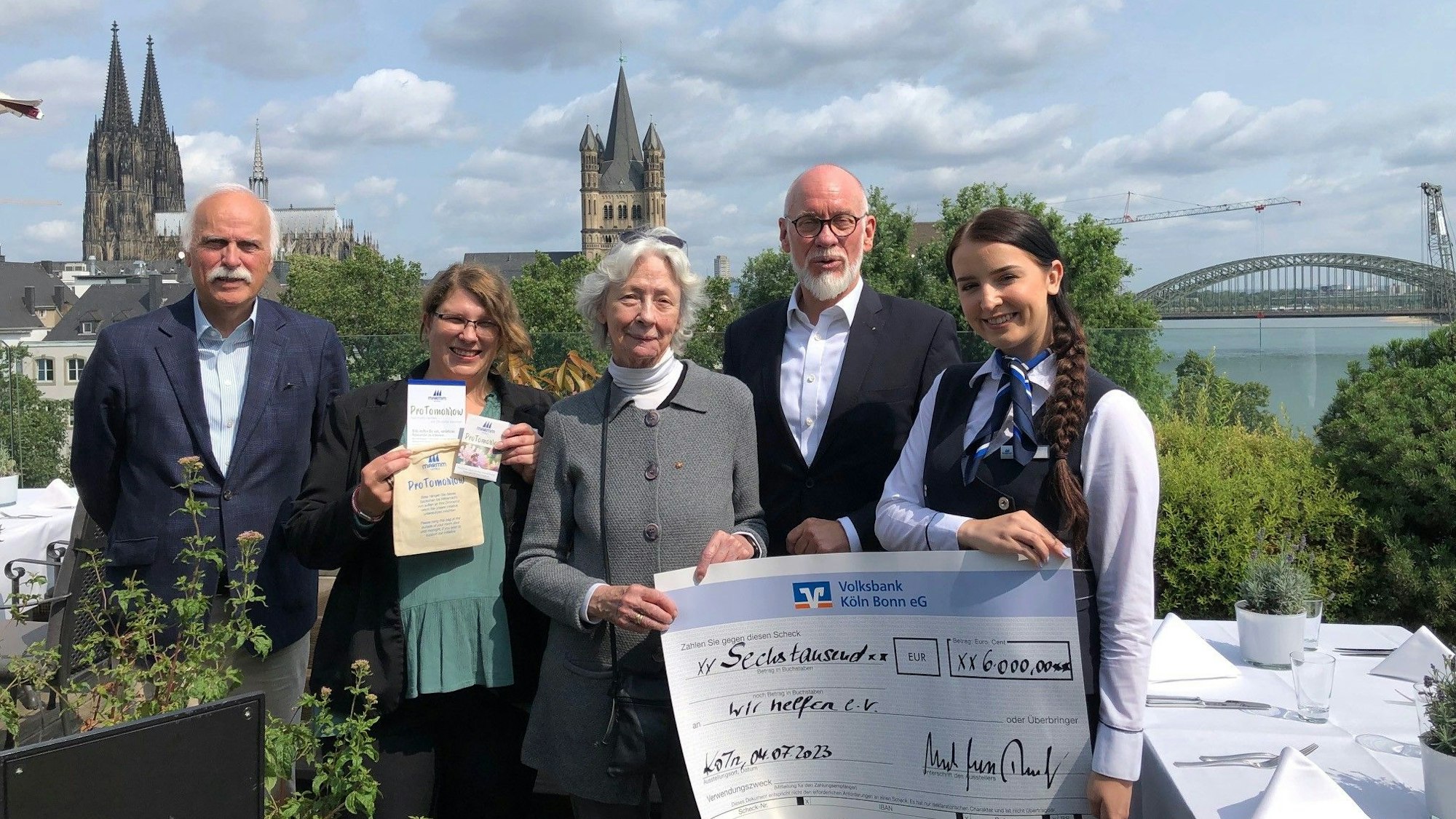Karl-Heinz Goßmann und Hedwig Neven DuMont von „wir helfen“ nehmen auf der Terrasse des „Bellevue“ mit Blick auf den Dom von einer Maritim-Hotel-Delegation einen symbolischen Scheck in Höhe von 6000 Euro entgegen.