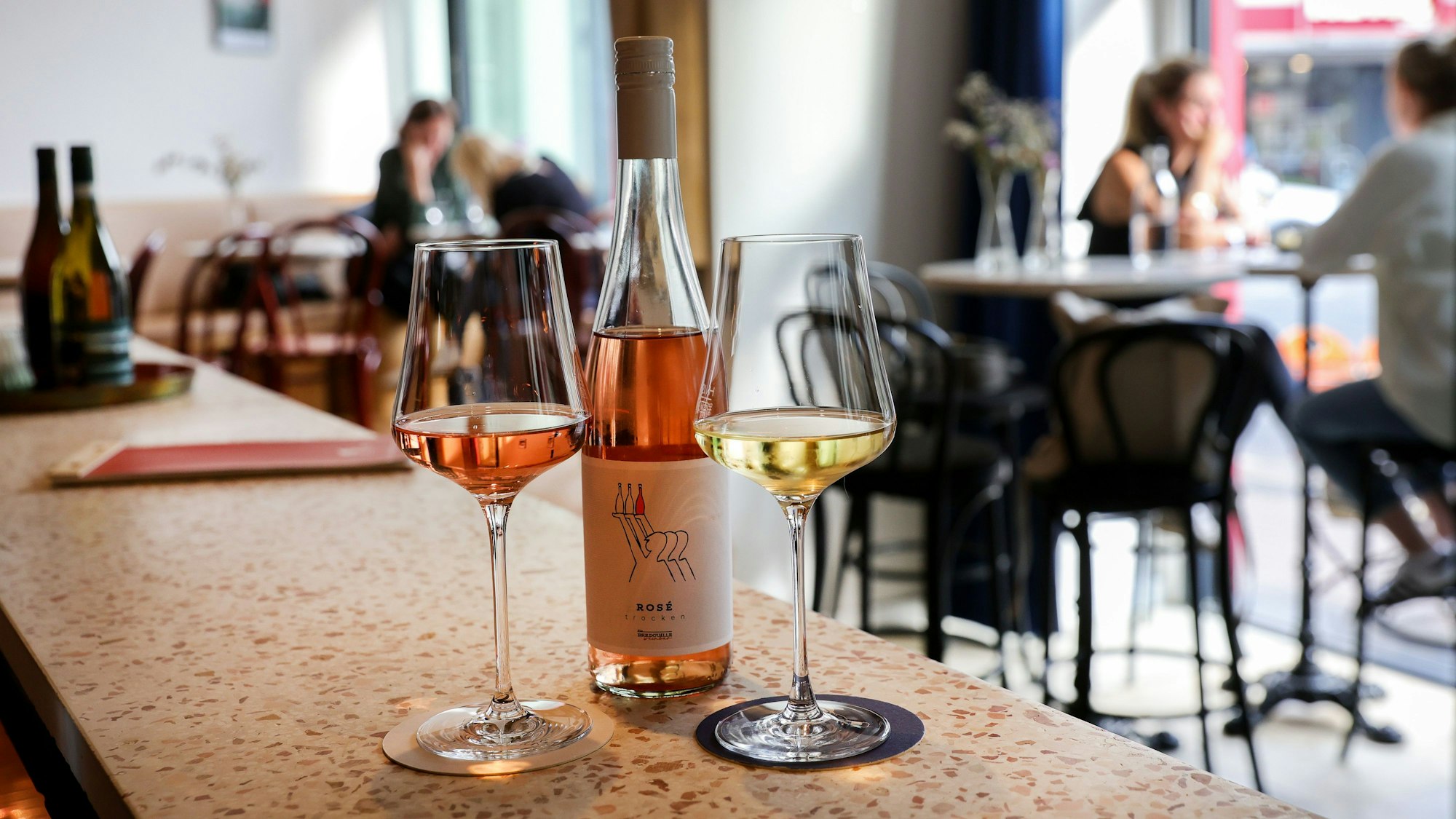 03.07.2023, Köln: Die Weinbar Bedrouille: Auf der Theke steht eine Flasche Wein mit Gläsern, im Hintergund sitzen Menschen an Tischen.