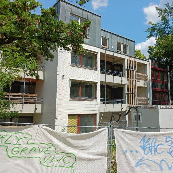 Ein Bauzaun steht vor einem mehrstöckigen Gebäude, das von Bäumen gesäumt wird.