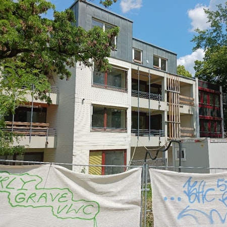 Ein Bauzaun steht vor einem mehrstöckigen Gebäude, das von Bäumen gesäumt wird.