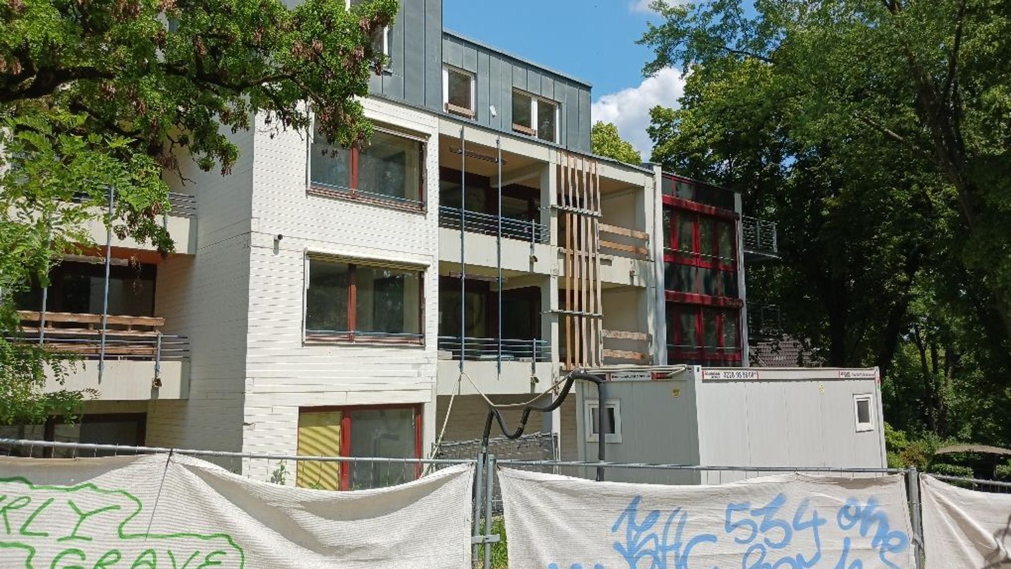 Ein Bauzaun steht vor einem mehrstöckigen Gebäude, das von Bäumen gesäumt wird.