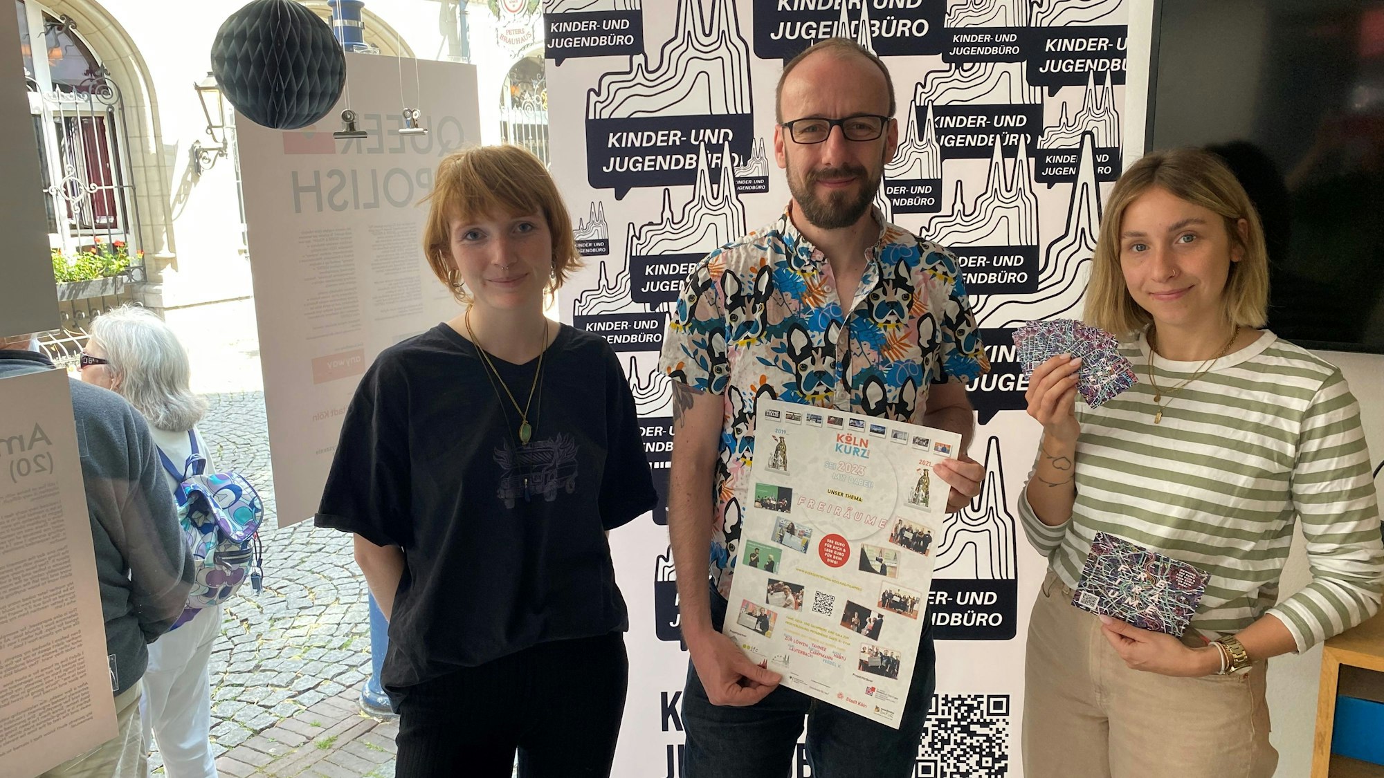 Auf dem Bild stehen drei Leute, die ein Plakat und Flyer des diesjährigen Jugendfilmpreis hochhalten.