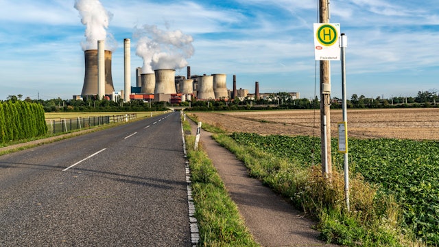Eine Bushaltestelle an einer Landstraße. Im Hintergrund ist ein Kohlekraftwerk mit dampfenden Türmen zu sehen.