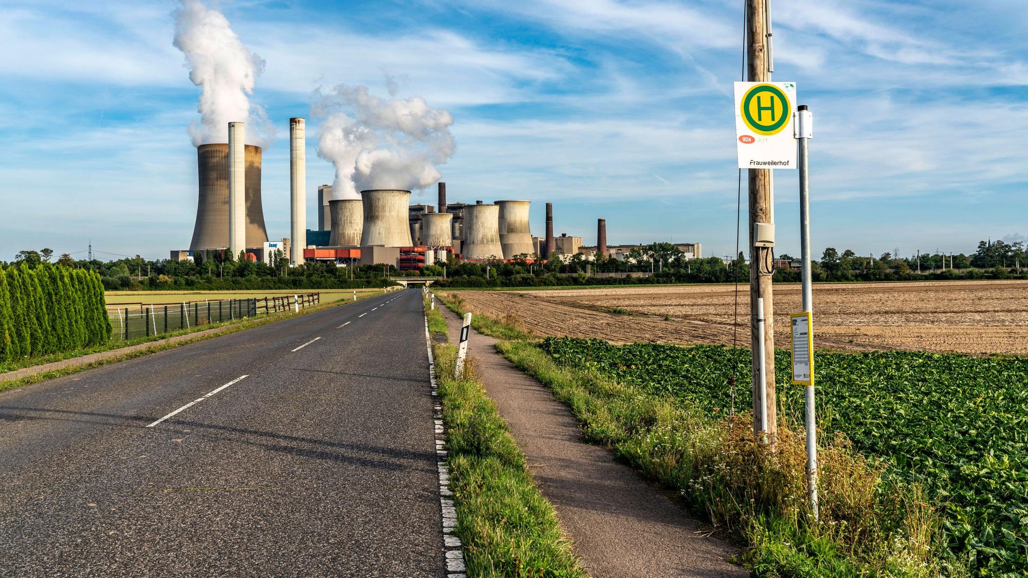 Eine Bushaltestelle an einer Landstraße. Im Hintergrund ist ein Kohlekraftwerk mit dampfenden Türmen zu sehen.