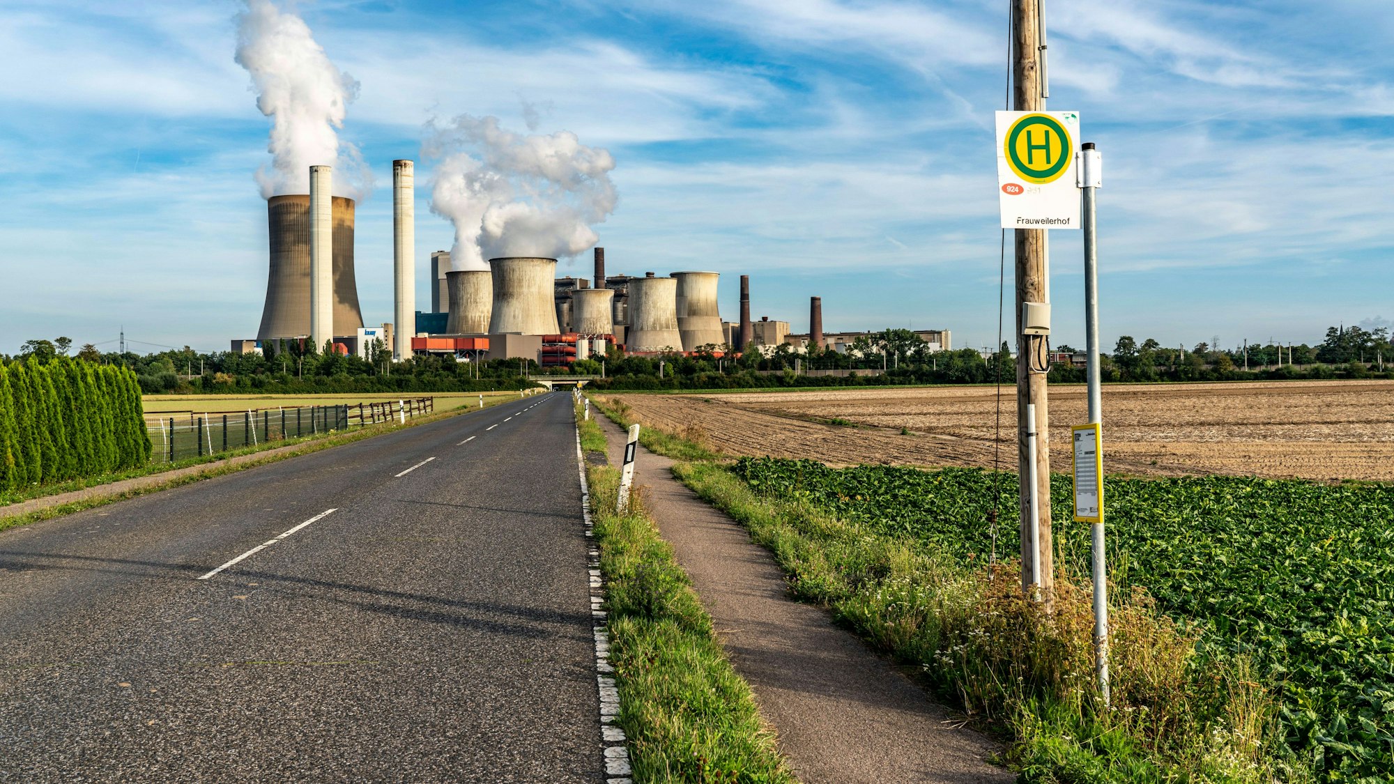 Eine Bushaltestelle an einer Landstraße. Im Hintergrund ist ein Kohlekraftwerk mit dampfenden Türmen zu sehen.