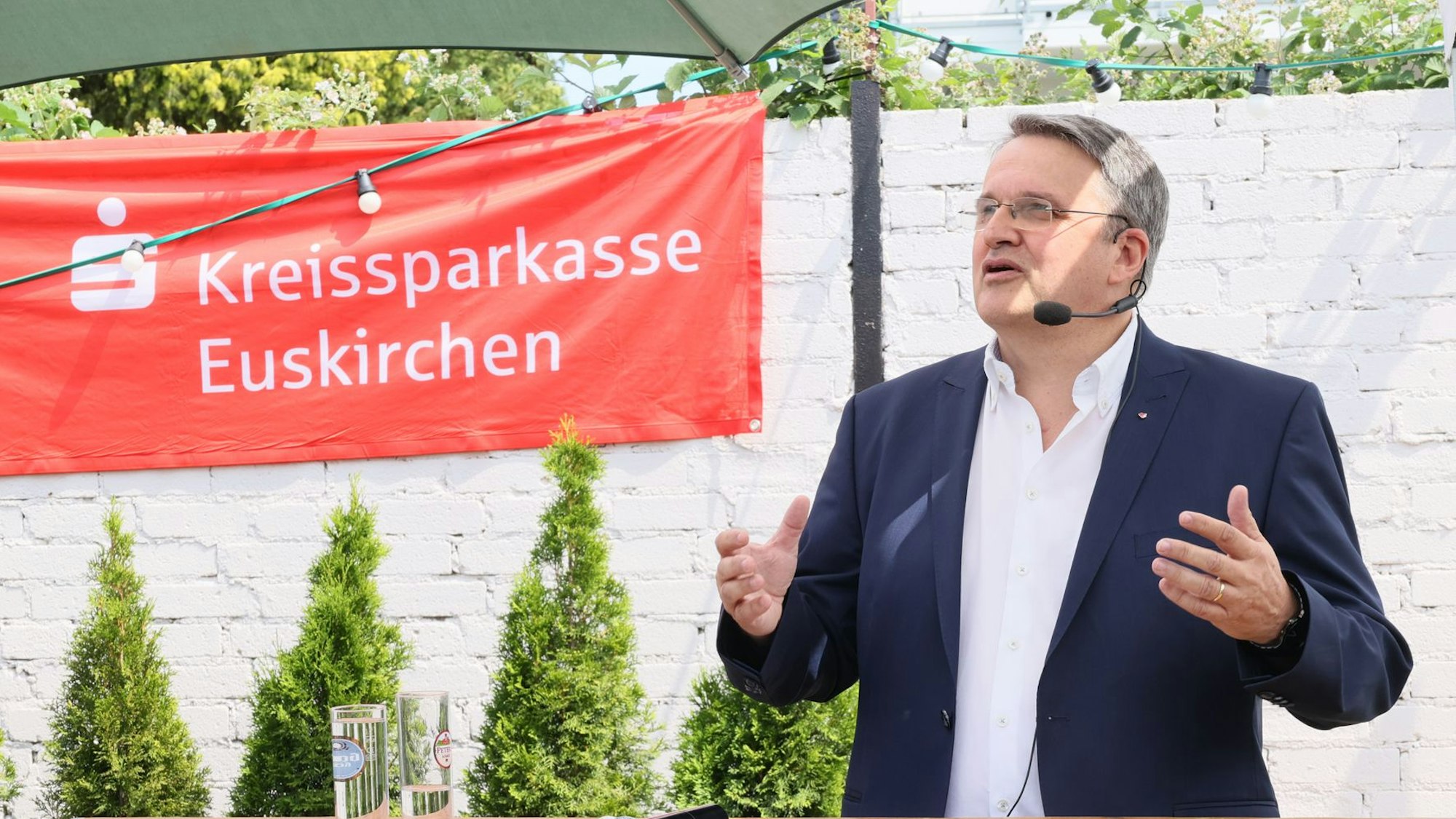 Udo Becker moderiert mit einem Headset-Mikrofon. Er trägt einen Anzug.