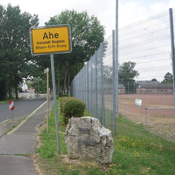 Das Bild zeigt das Ortsschild von Ahe, daneben eine Straße und den Aschenplatz des Dorfs.