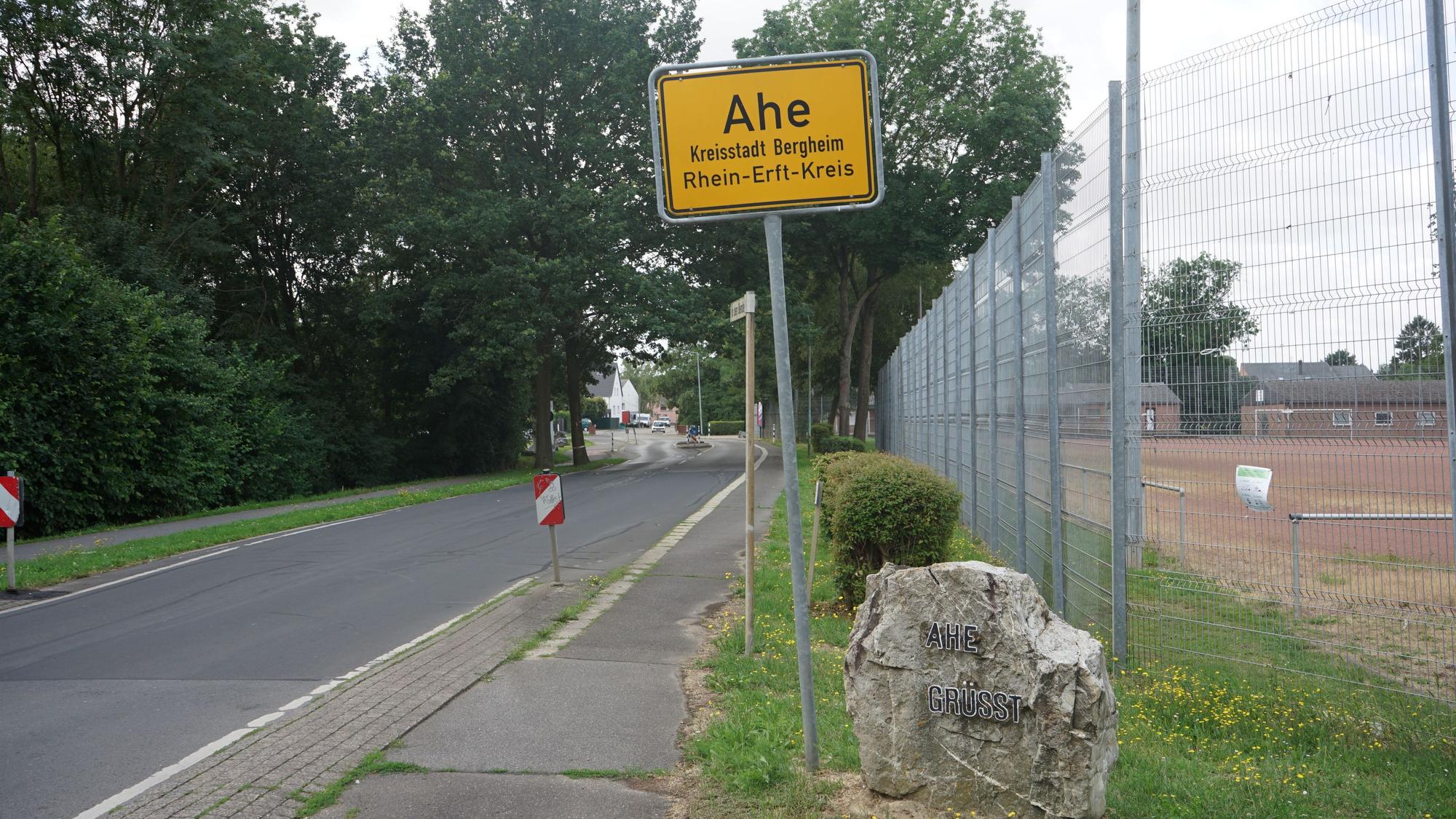 Das Bild zeigt das Ortsschild von Ahe, daneben eine Straße und den Aschenplatz des Dorfs.