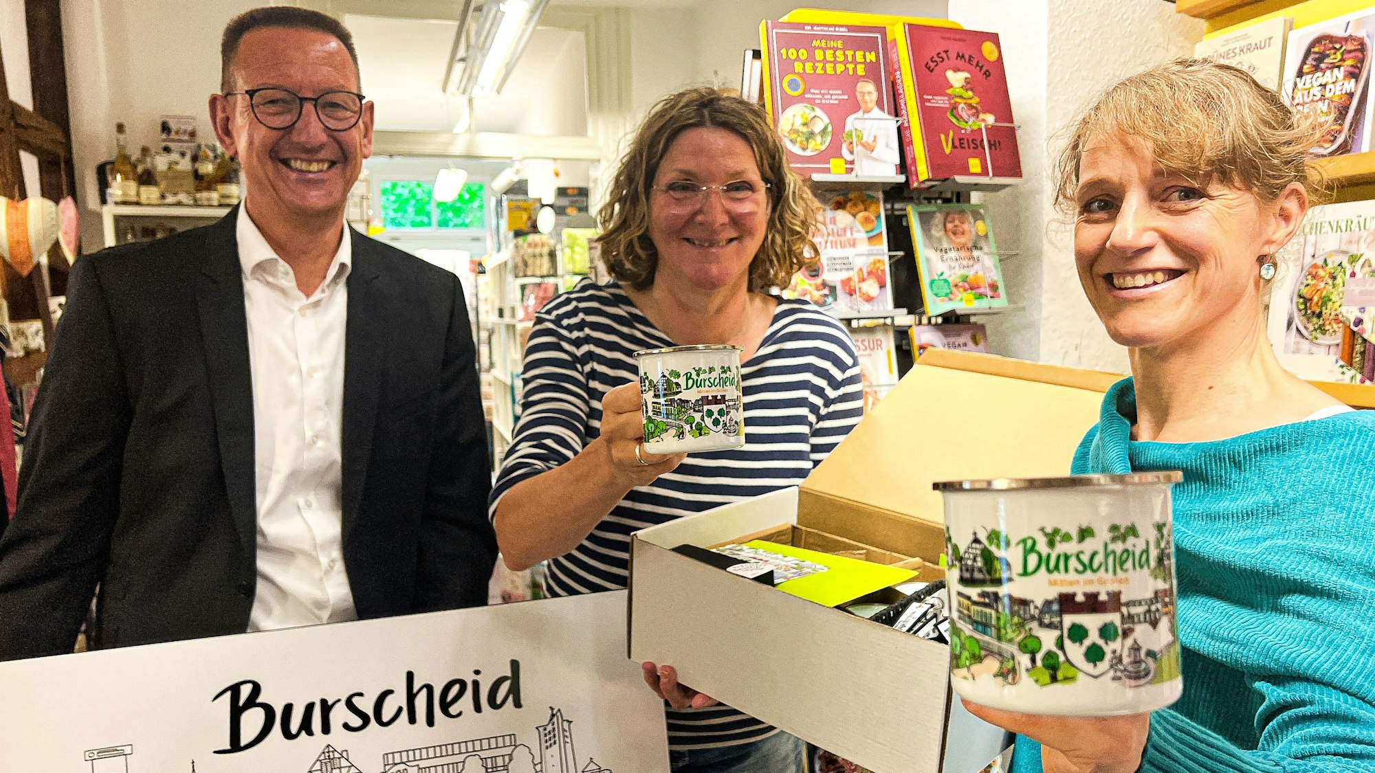 Neues Merchandise für Burscheid: Bürgermeister Drik Runge (v.l.) mit der Burscheid-Skyline, Buchhändlerin Ute Hentschel und Illustratorin Myria Stricker mit Burscheid-Emailletassen.
