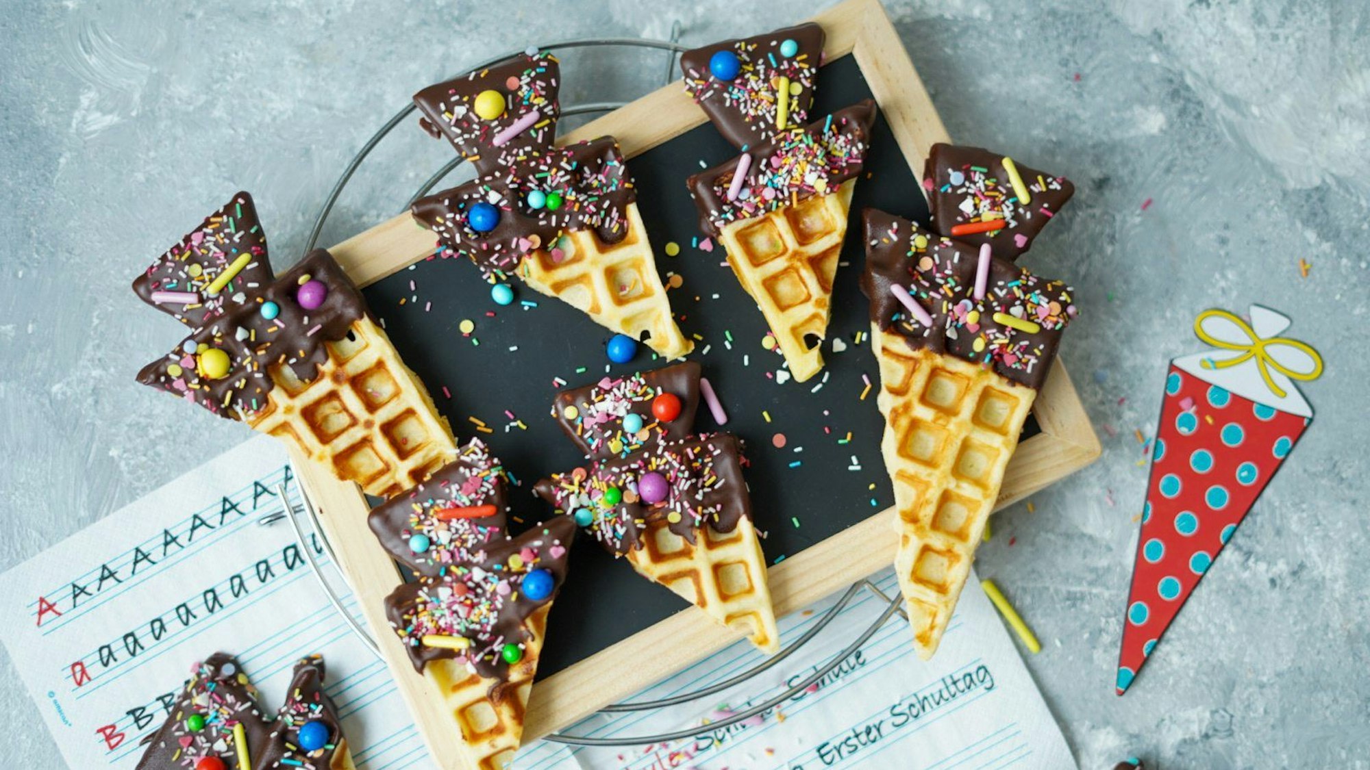 Waffeln in Form einer Schultüte liegen auf einem Tisch. Die Waffeln sind mit Schokolade und bunten Zuckerperlen verziert.