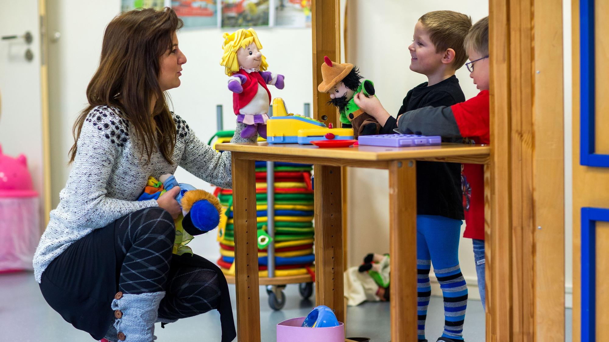Eine Frau und zwei Kinder spielen mit Handpuppen. Die Frau hockt auf dem Boden vor den Kindern.